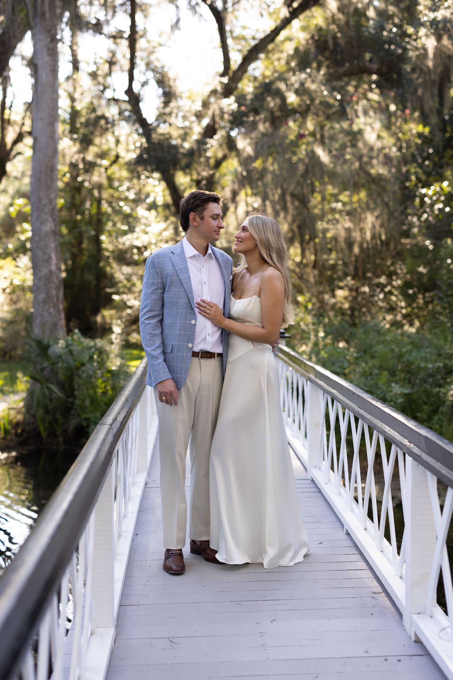 Couple in wedding attire sharing a romantic moment on a white wooden bridge surrounded by lush green trees.