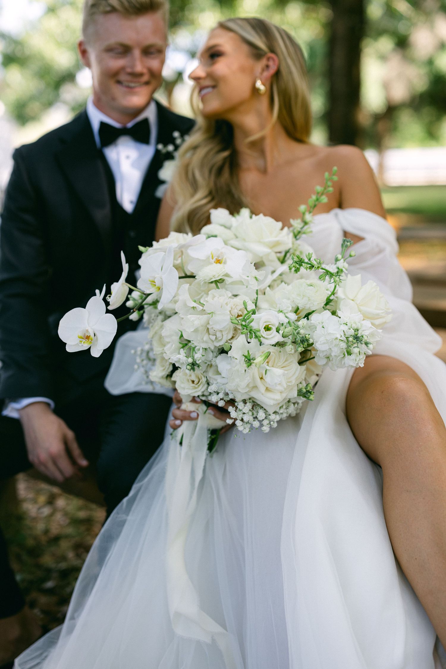 Bride in white off-shoulder gown holds lush white rose bouquet beside smiling groom in black tuxedo outdoors.