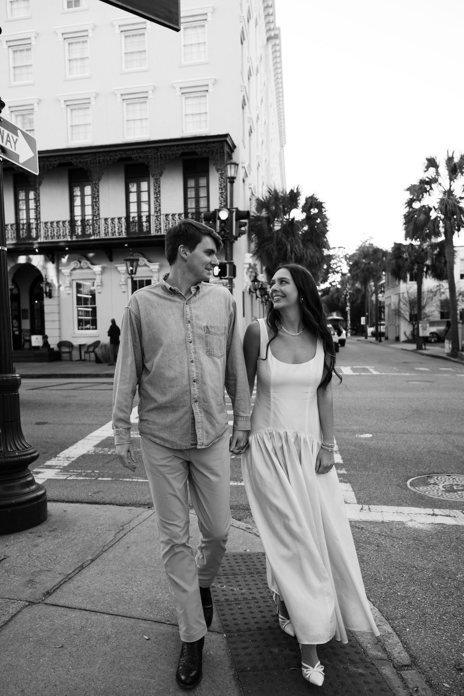 Black and white photo of a couple holding hands and smiling while walking across a city street crosswalk.