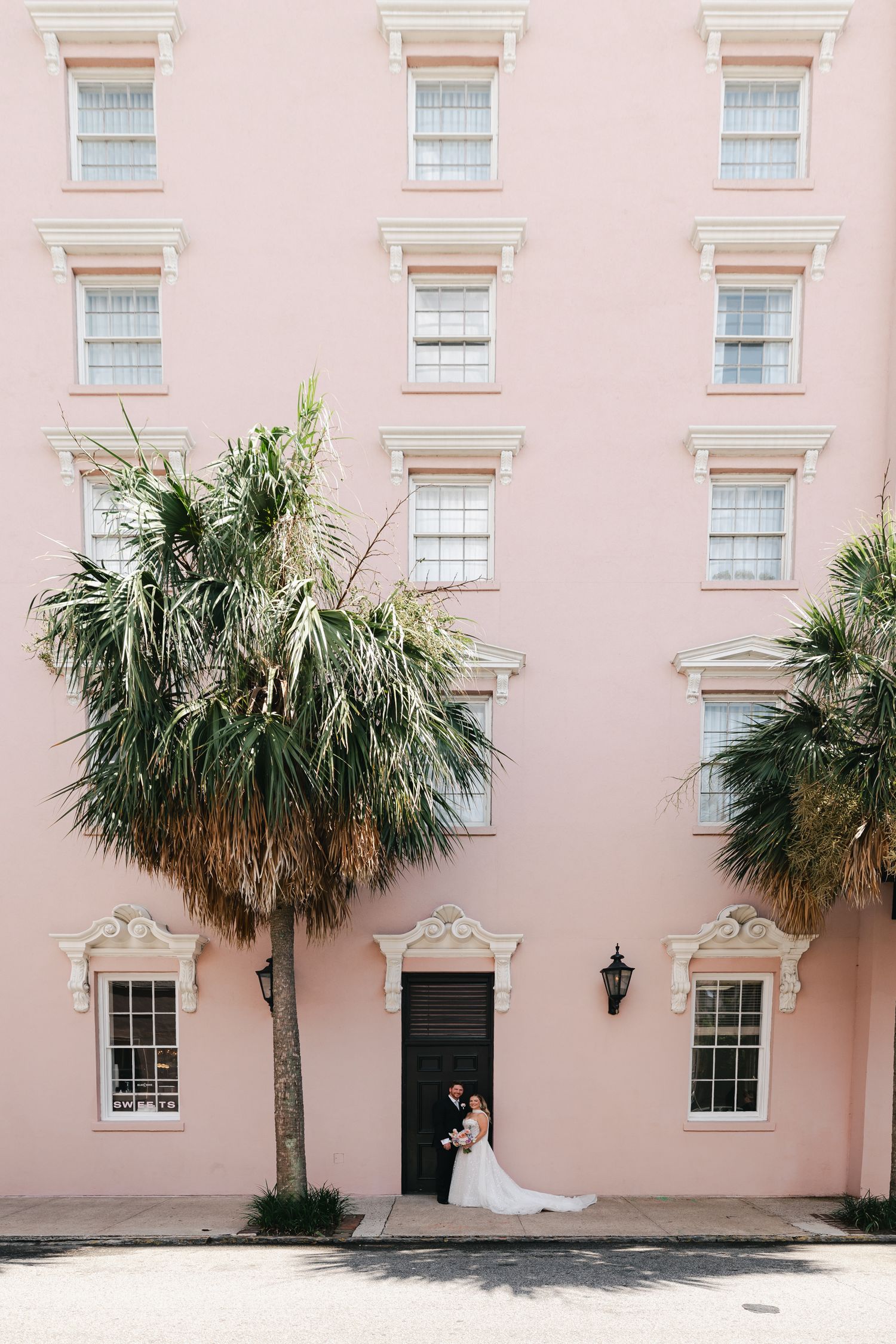 Bride and groom embrace in a black doorway of a pink building with palm trees in Charleston, SC.