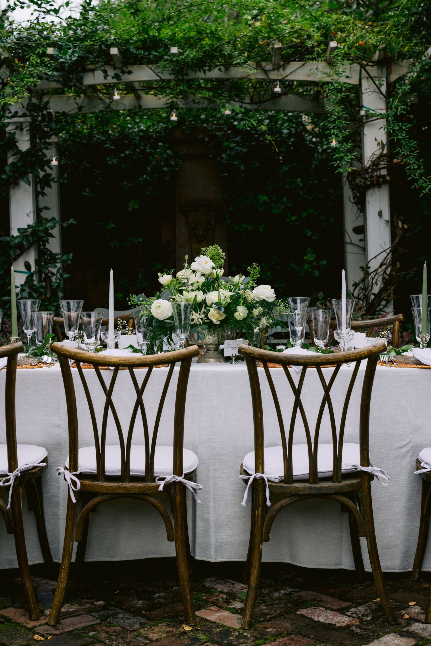Elegant outdoor wedding reception table with white linens, floral centerpiece, and rustic wooden chairs under a pergola.