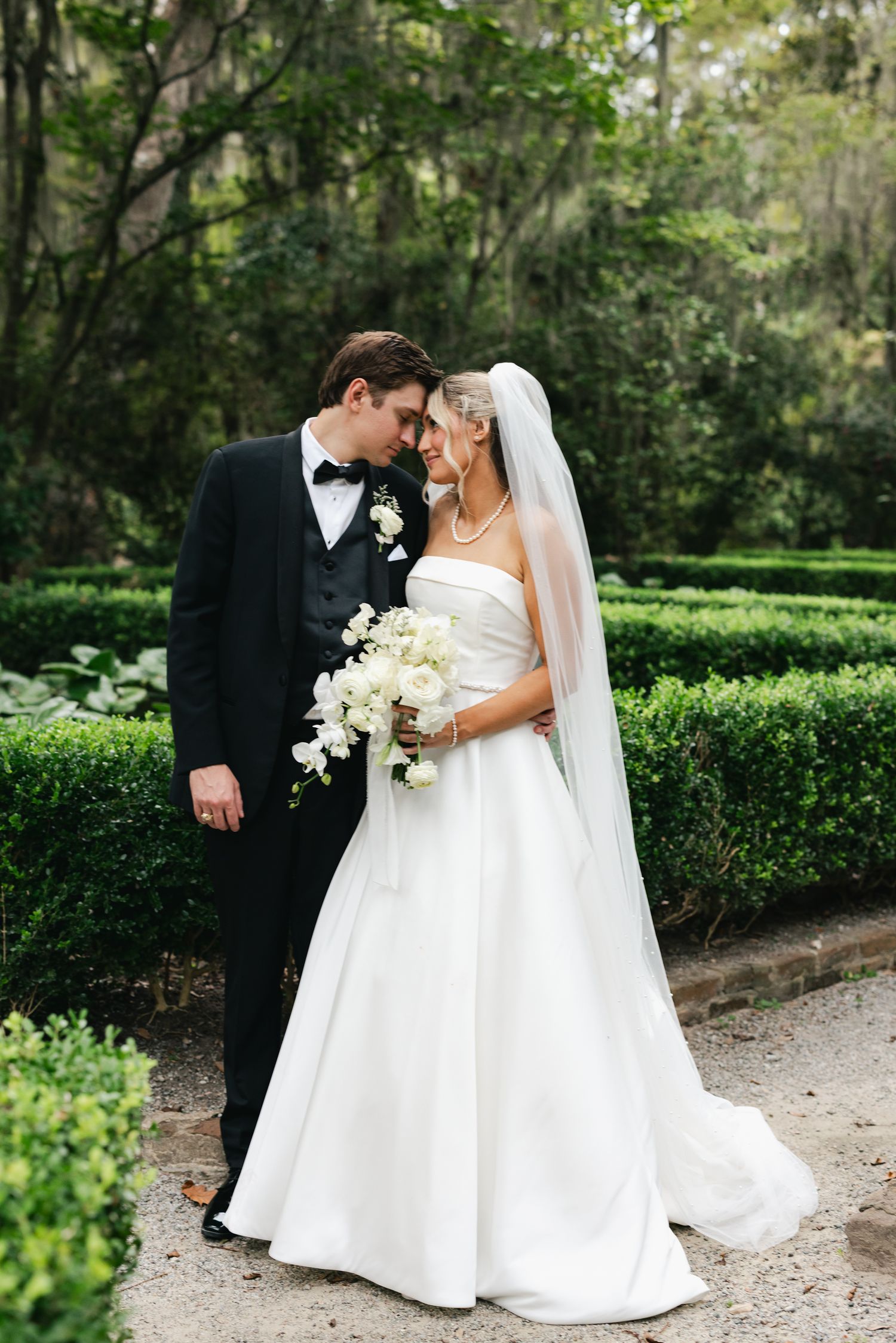 Bride in strapless ball gown and groom in black tuxedo sharing romantic moment in lush garden.