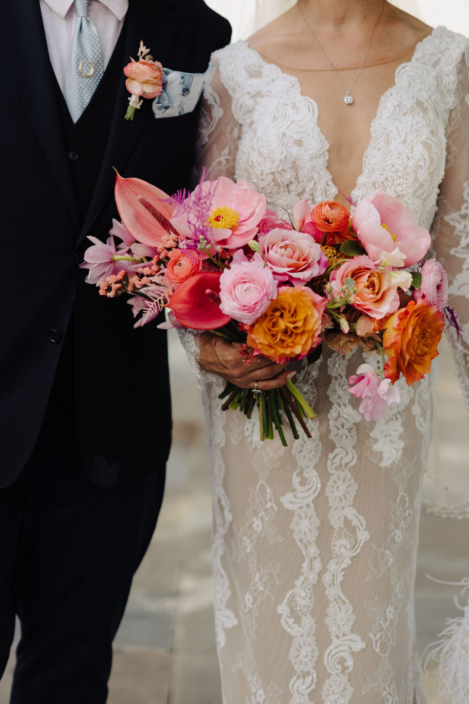 Bride in lace gown holds vibrant pink and orange wedding bouquet beside groom in navy suit with boutonniere.