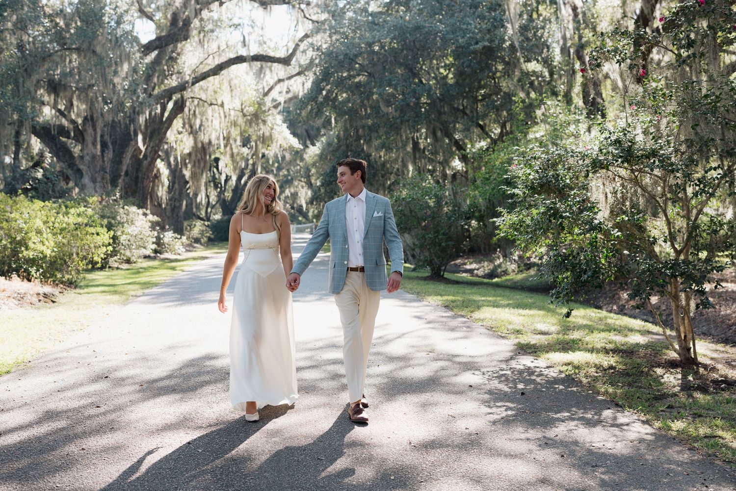 Bride and groom walk hand in hand down scenic path lined with Spanish moss-covered oak trees.