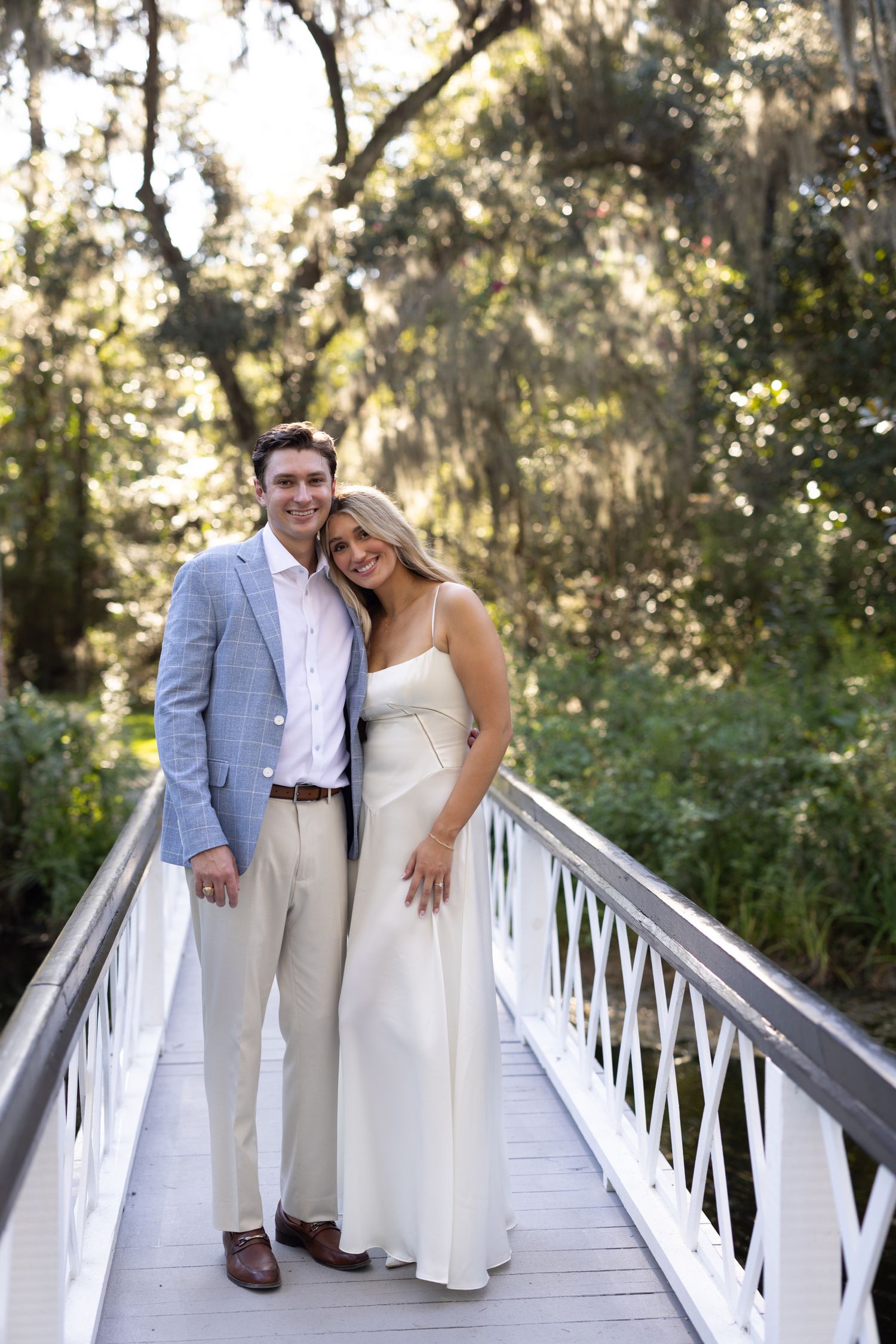 Couple in elegant attire posing on a white wooden bridge surrounded by moss-draped trees.