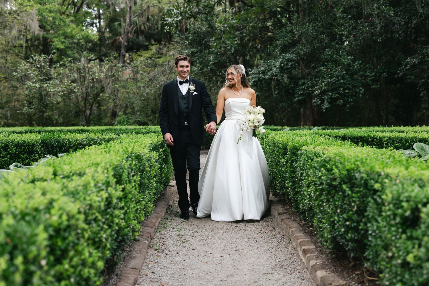 Bride and groom walk hand-in-hand through lush green hedge garden path on their wedding day.