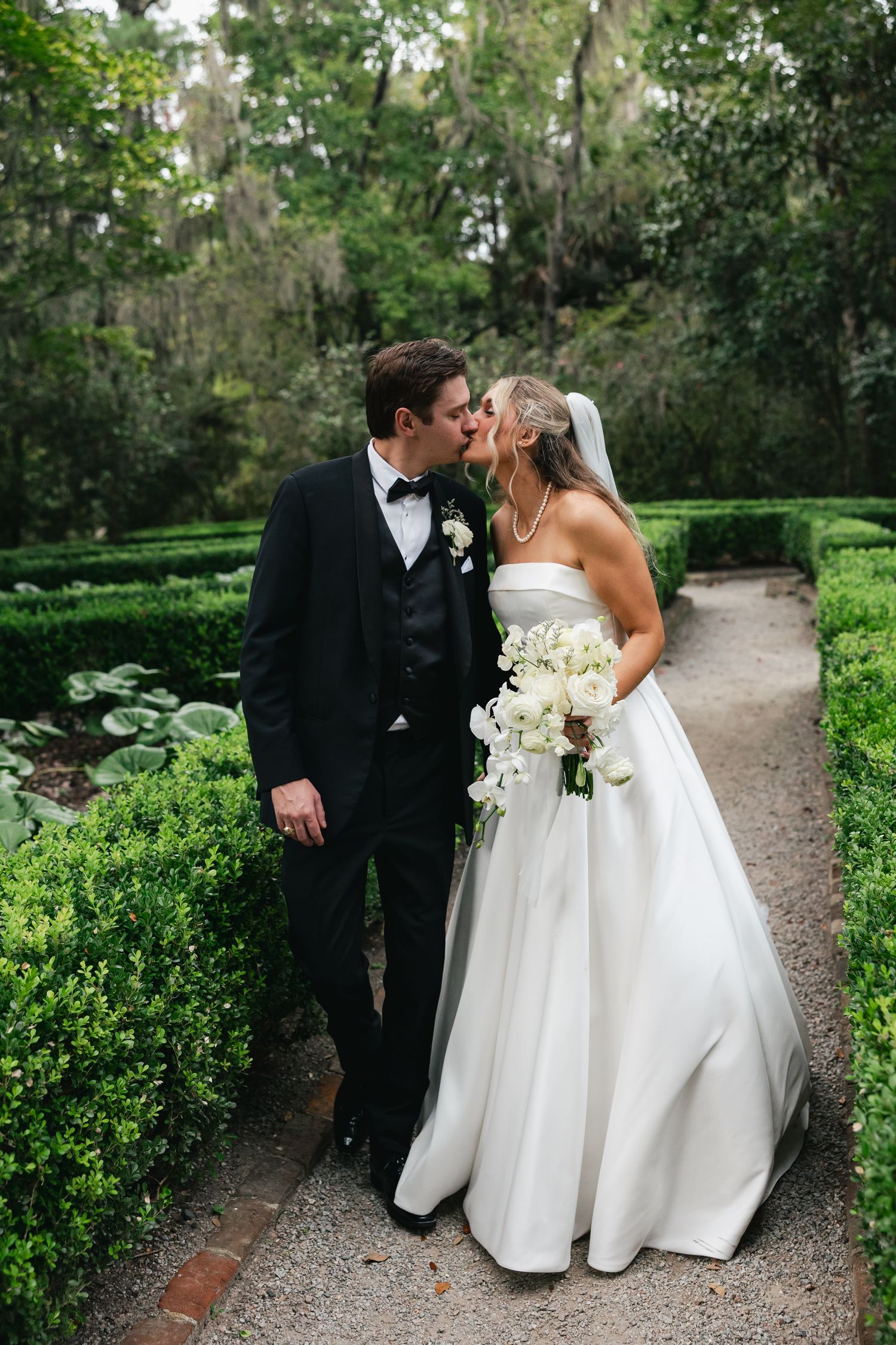 Bride and groom share a kiss in a lush garden pathway, she holds white florals bouquet, he wears black tuxedo.