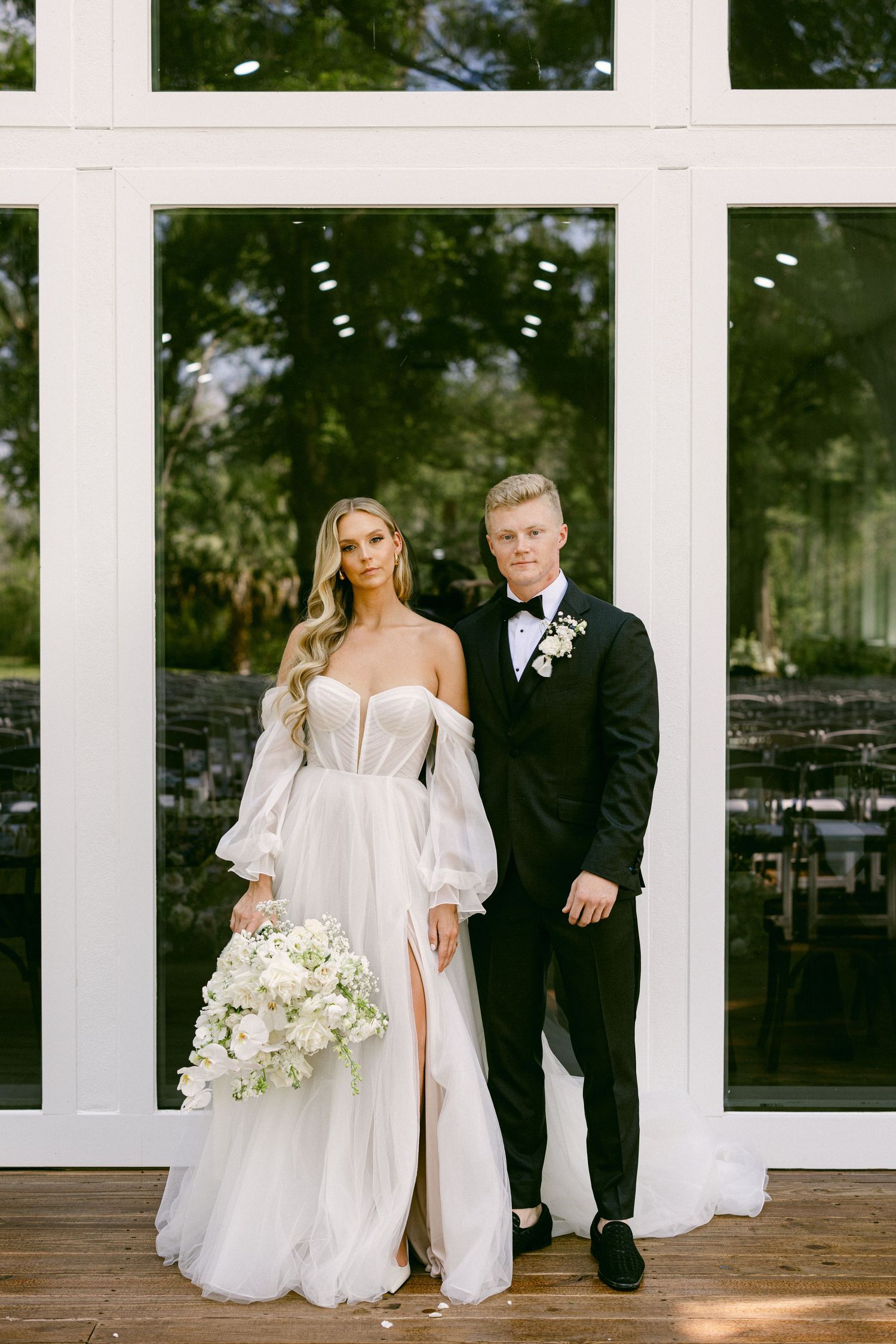 Bride in strapless tulle wedding gown holding white bouquet stands with groom in black tuxedo before large glass windows.