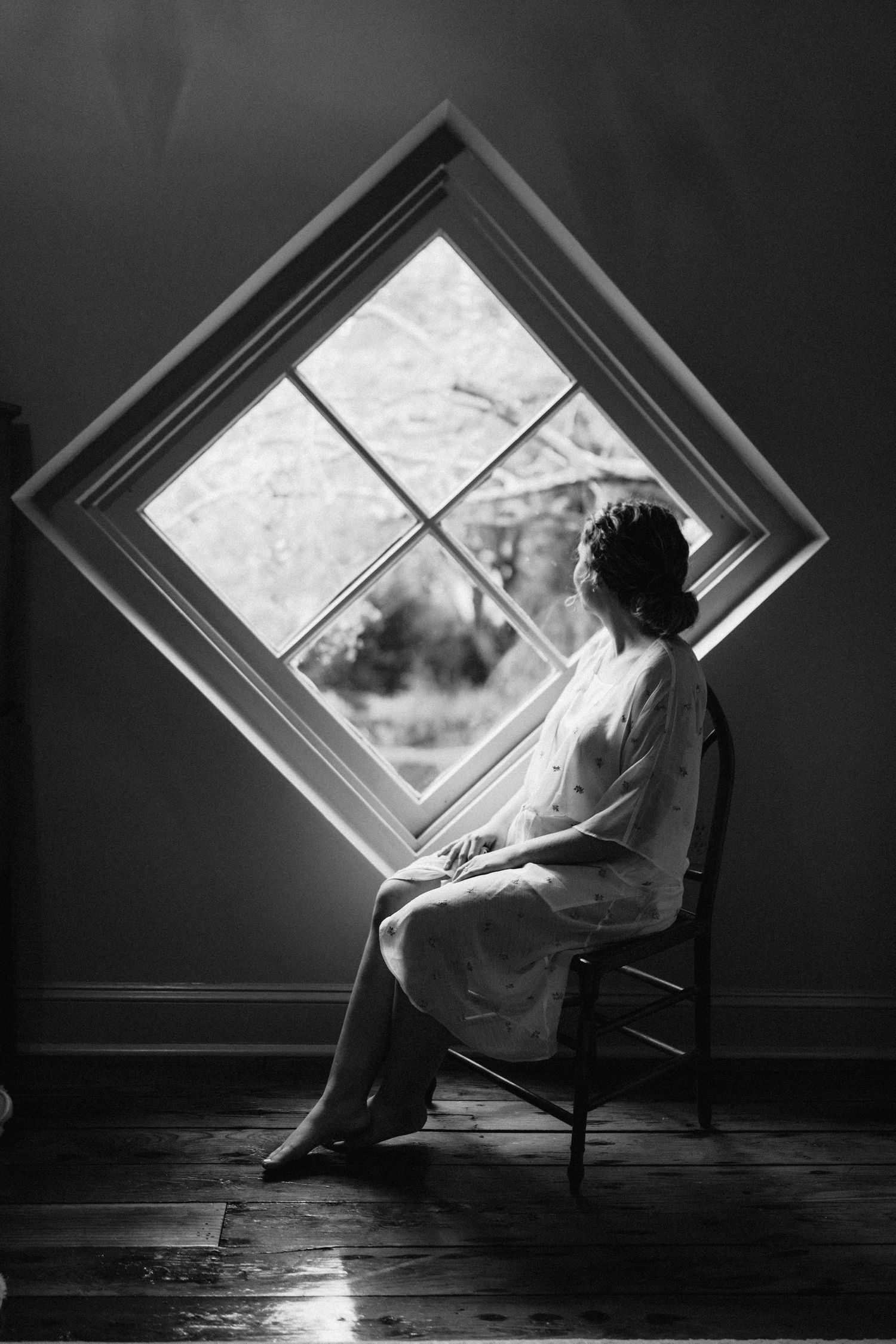 Black and white photo of bride sitting by diamond-shaped window, silhouetted in soft natural light.