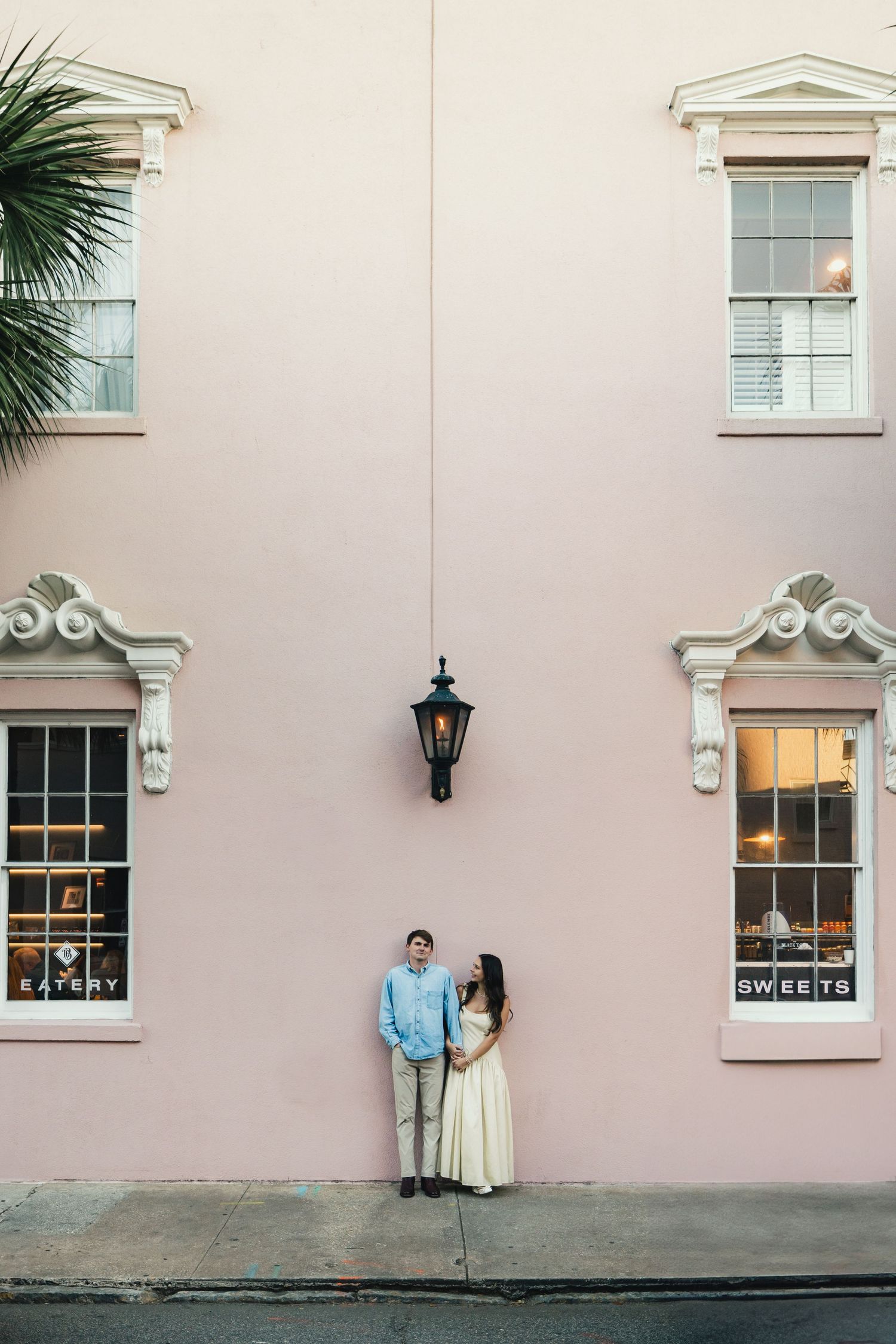 Couple poses in front of a pink building with a lantern, flanked by windows showing Eatery and Sweets signs.