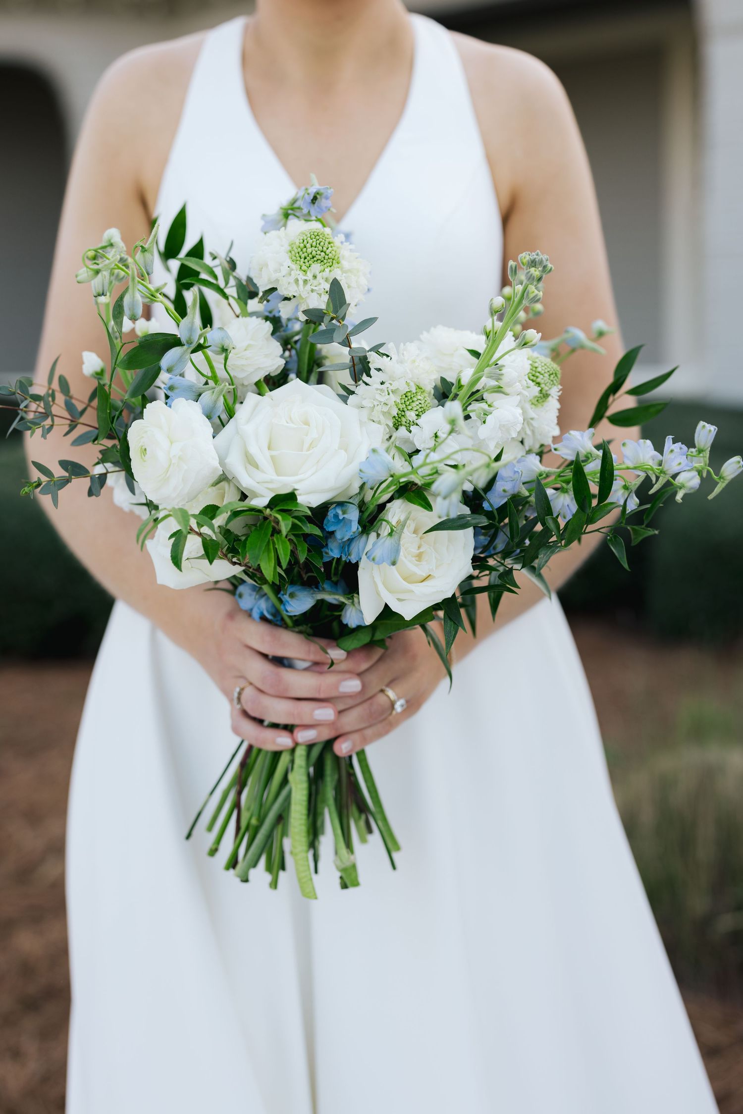Bride in white halter gown holding elegant bouquet of white roses, blue flowers, and greenery.
