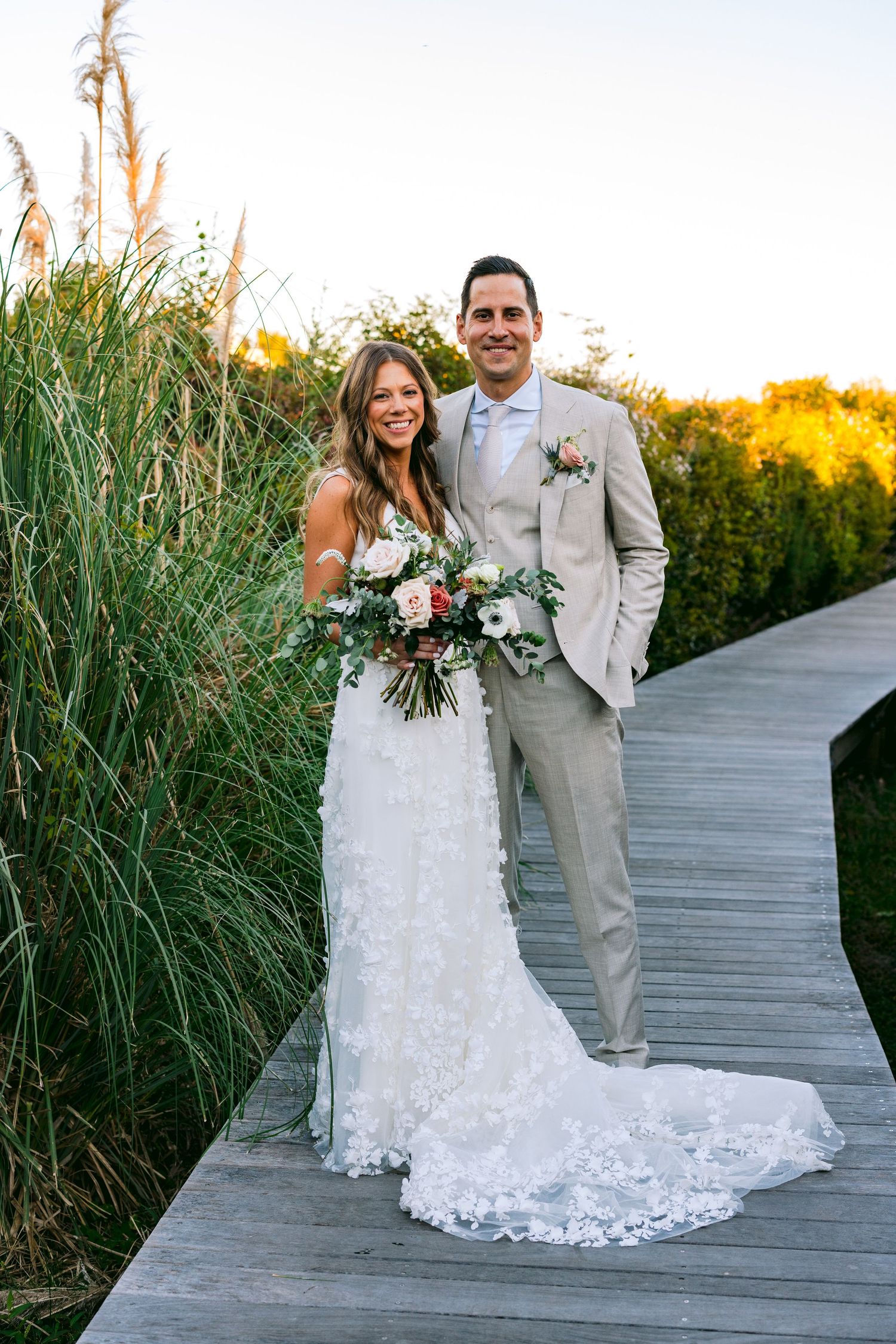 Bride and groom posing on wooden boardwalk surrounded by tall grasses, bride holding floral bouquet in lace gown.