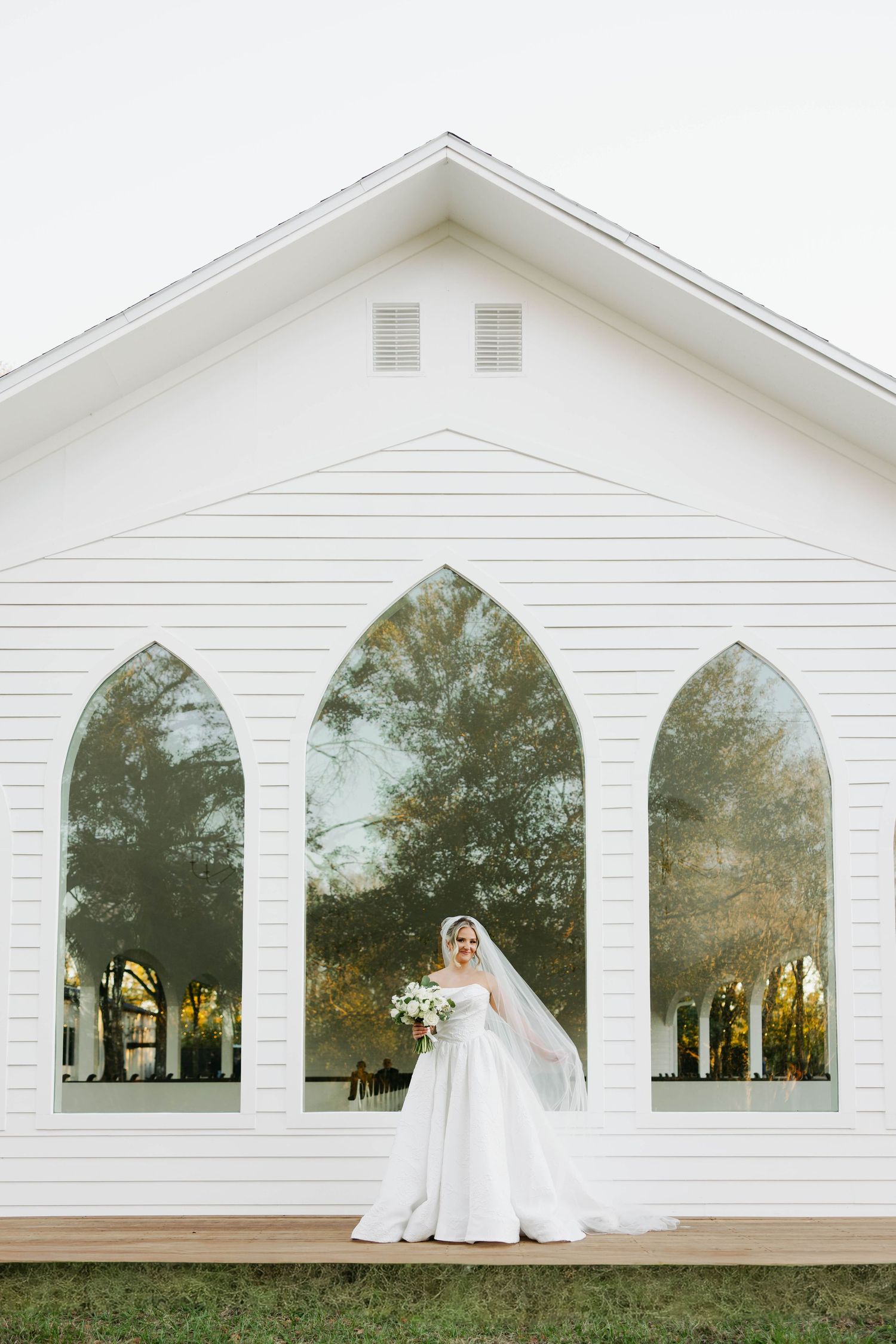 Bride in a white ball gown and veil stands before a white chapel with Gothic arch windows reflecting autumn trees.