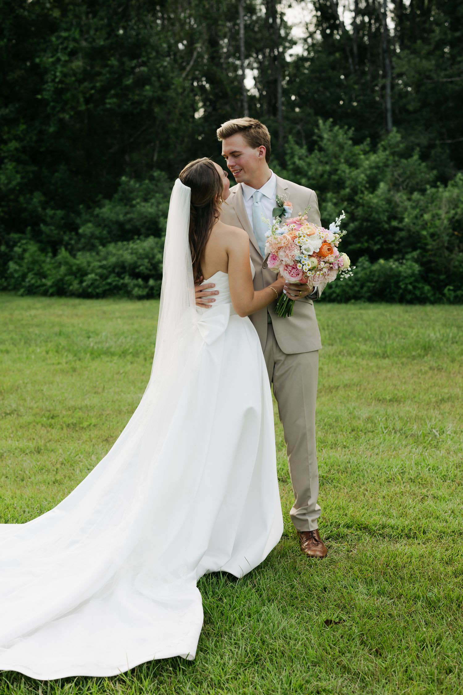 A bride in a white gown and veil holds a floral bouquet while embracing a groom in a tan suit on a green lawn.