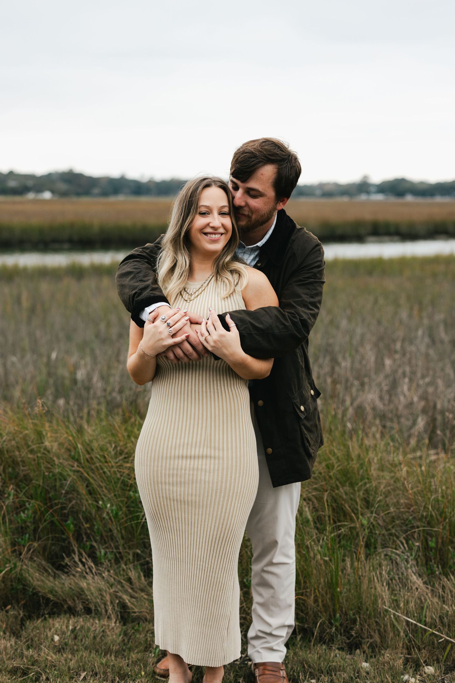 A couple shares a tender embrace in a marsh field, the pregnant woman in a cream ribbed dress held lovingly from behind by her partner.
