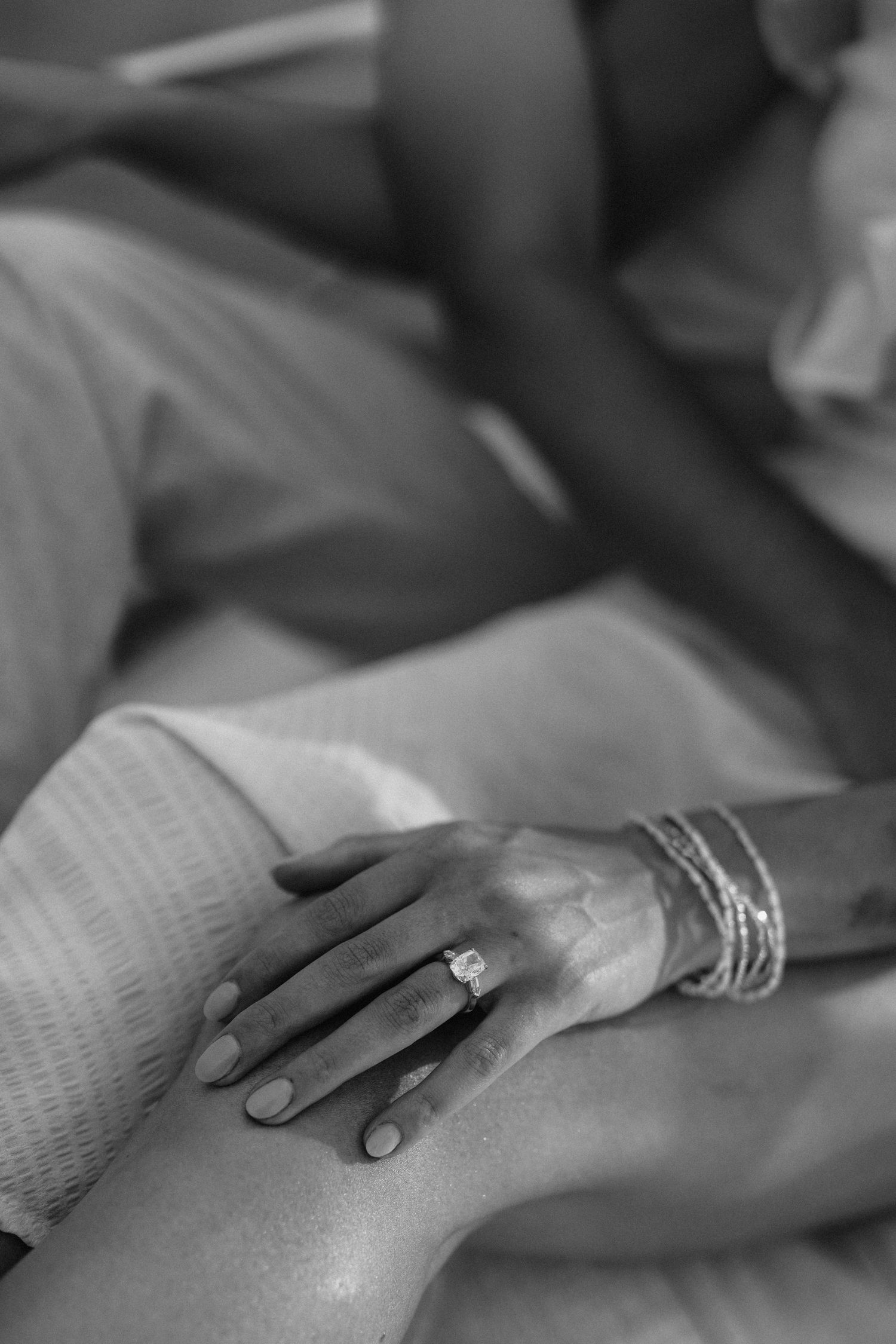 Black and white photo of a woman's hand with engagement ring and bracelets resting on a knee.