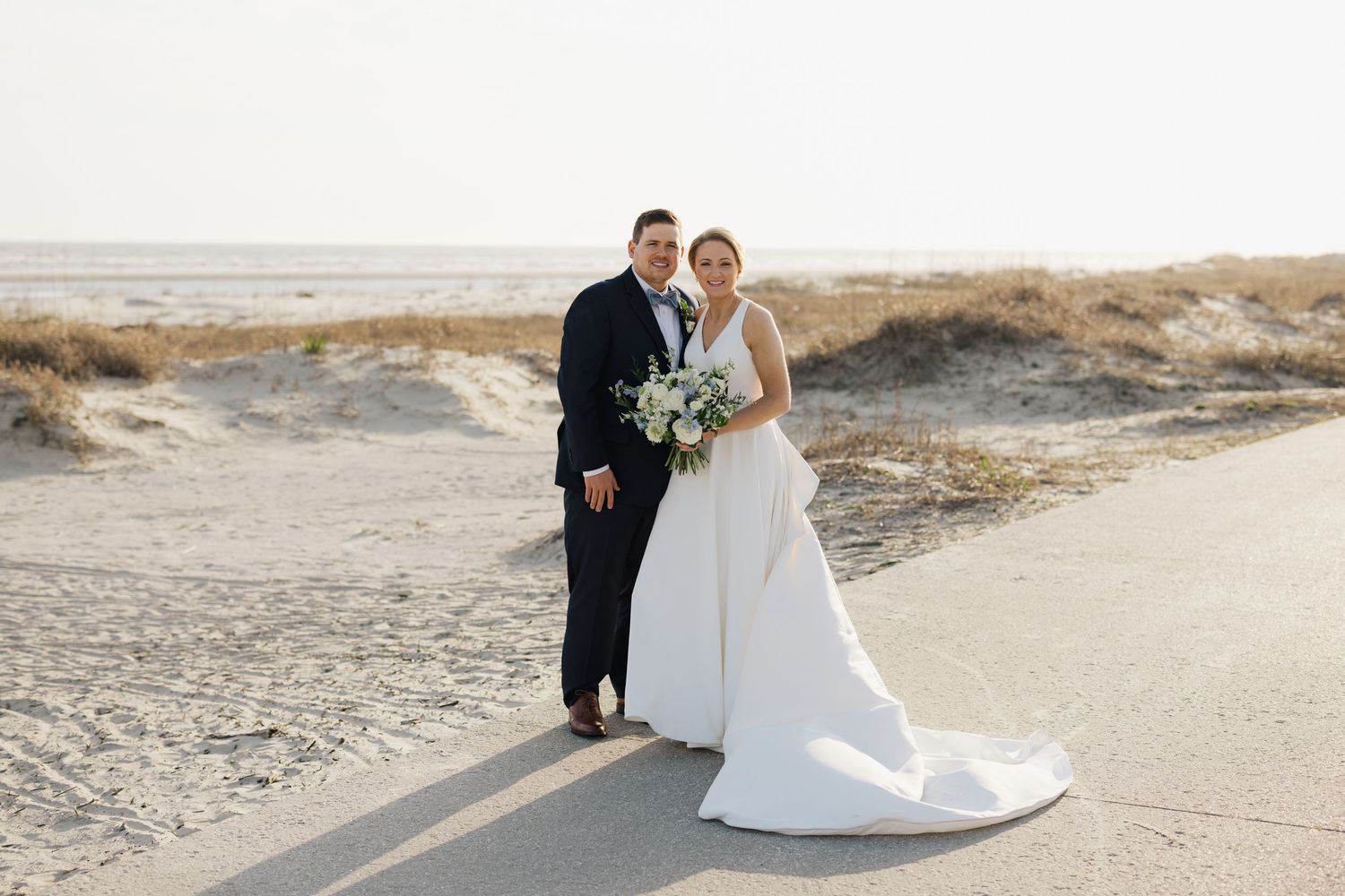 Bride and groom pose on a sandy beach path, she in a white gown with train, he in a dark suit, holding a bouquet.