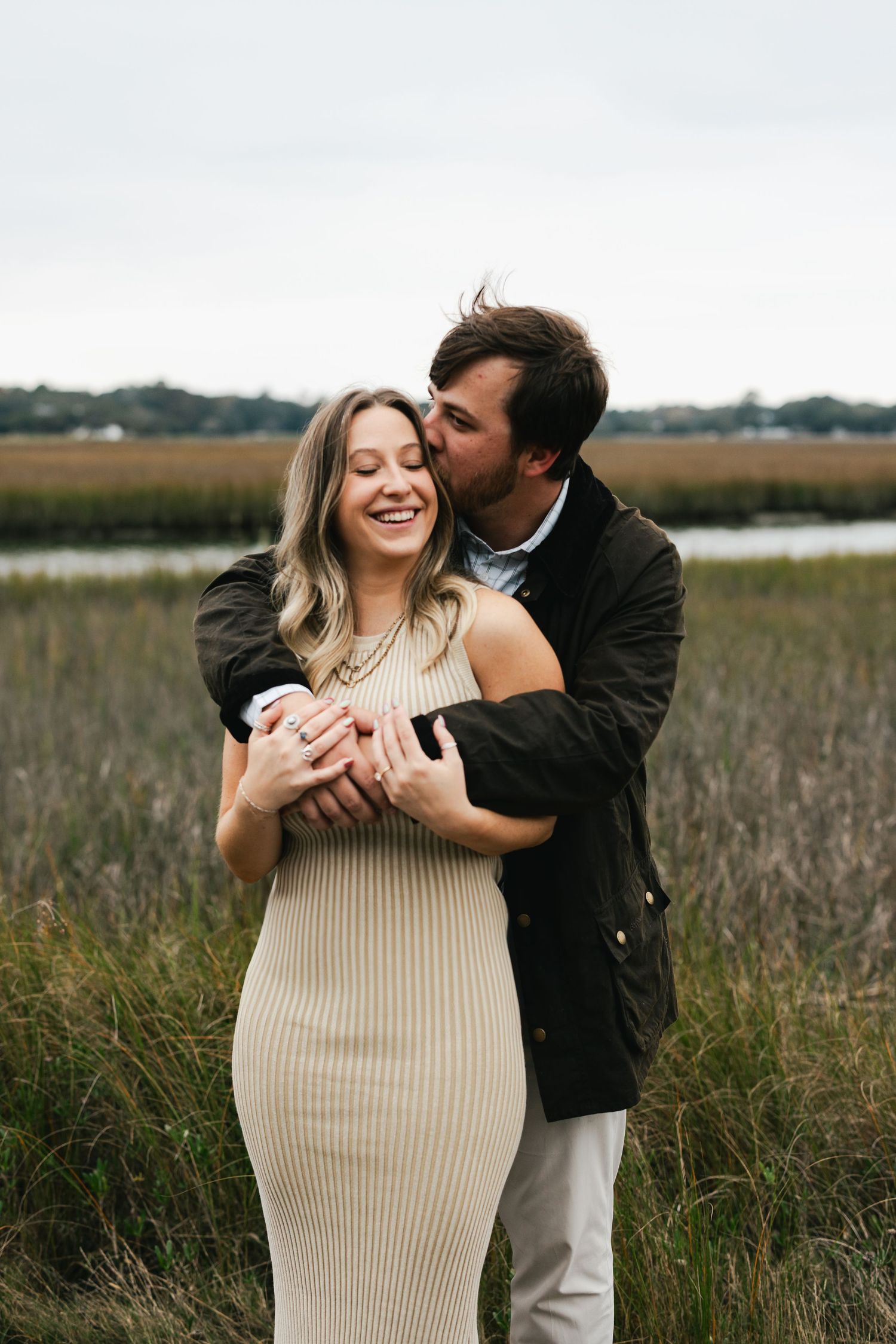 Man kissing laughing woman in beige ribbed dress near coastal marsh during engagement session.