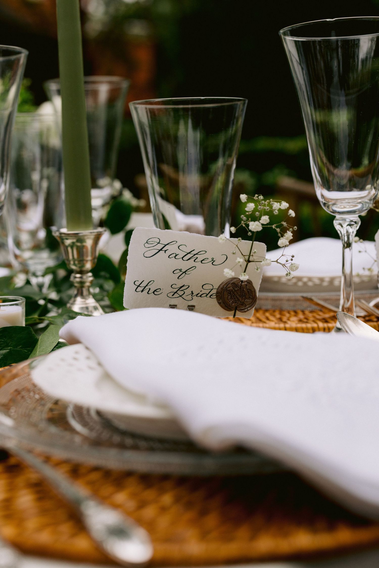Elegant wedding place setting with Father of the Bride card, white napkin, crystal glasses, and greenery on wicker charger.