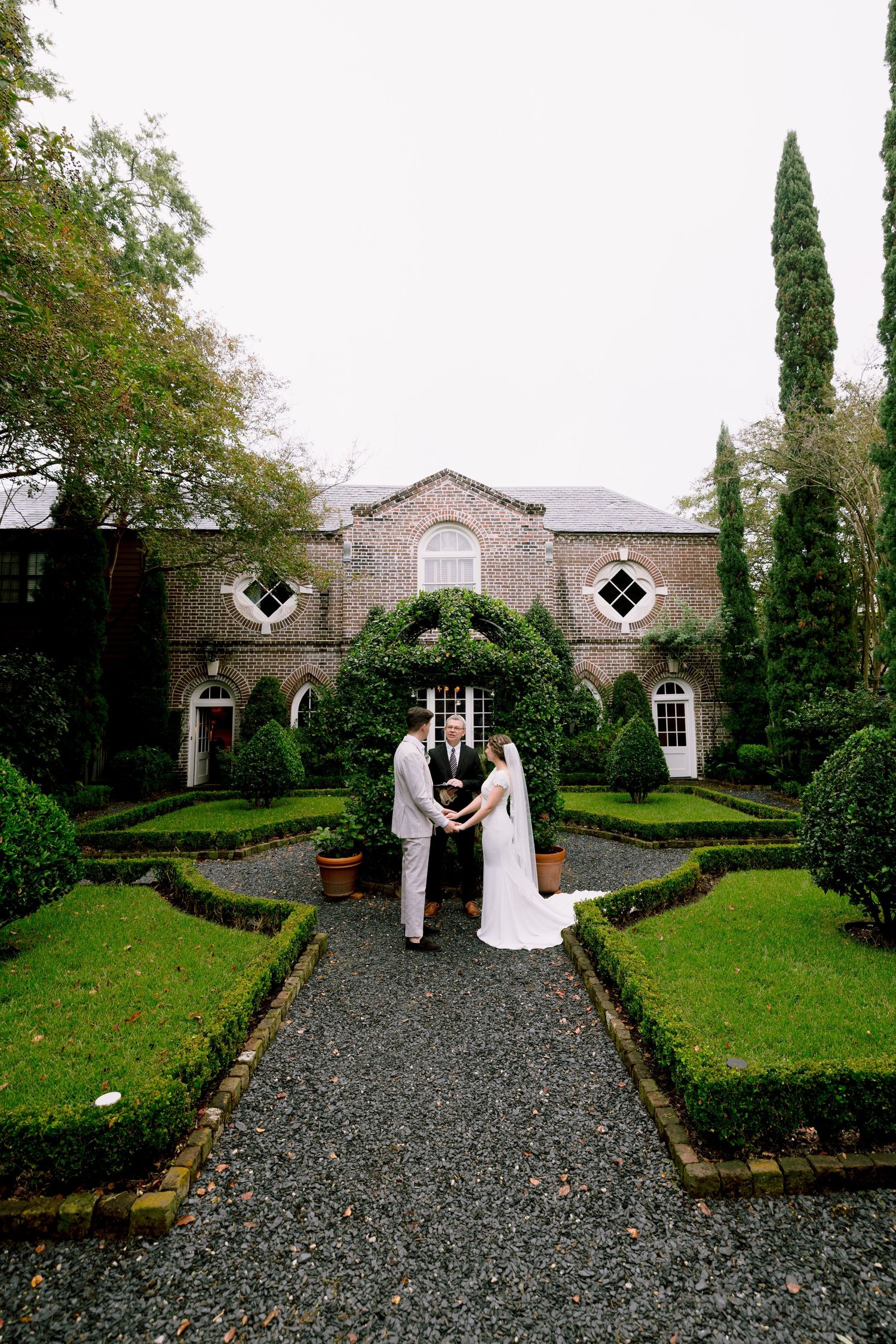 Bride and groom exchange vows in a formal garden with manicured hedges before an elegant brick manor.