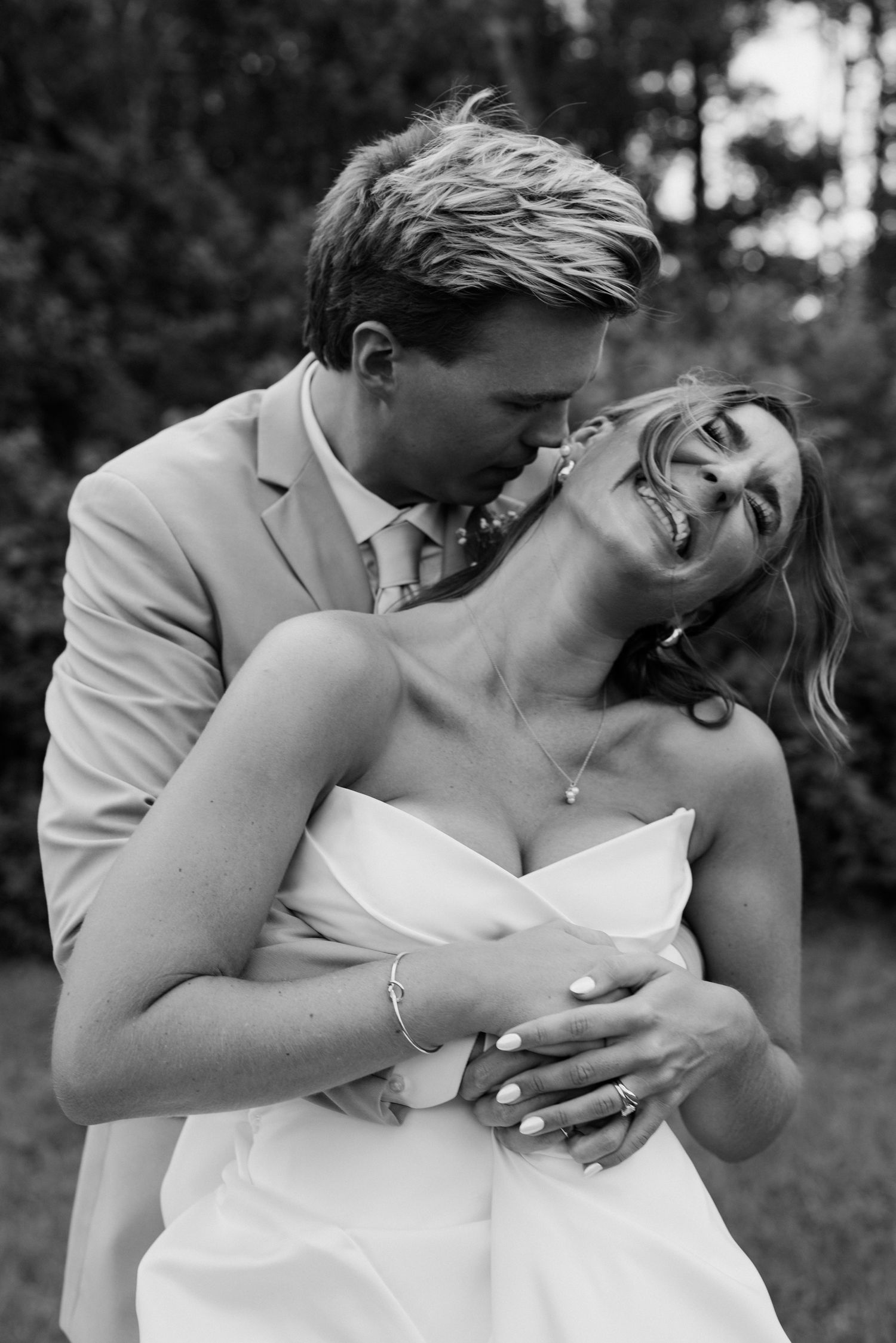 Black and white photo of groom in tan suit kissing laughing bride in strapless gown outdoors.