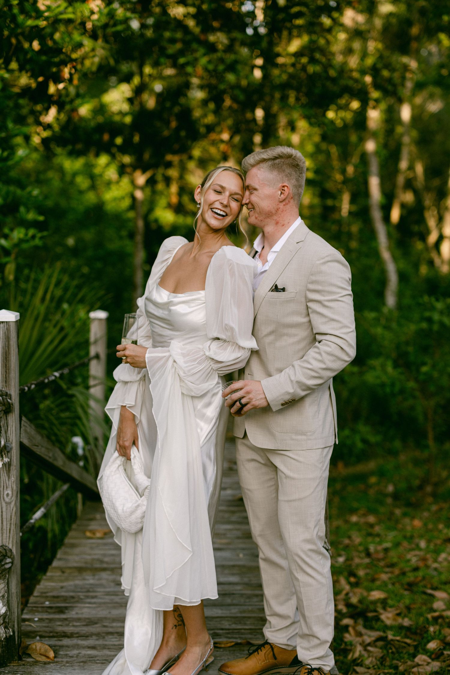 Laughing bride in white satin gown and groom in beige suit embrace on wooden bridge in forest.