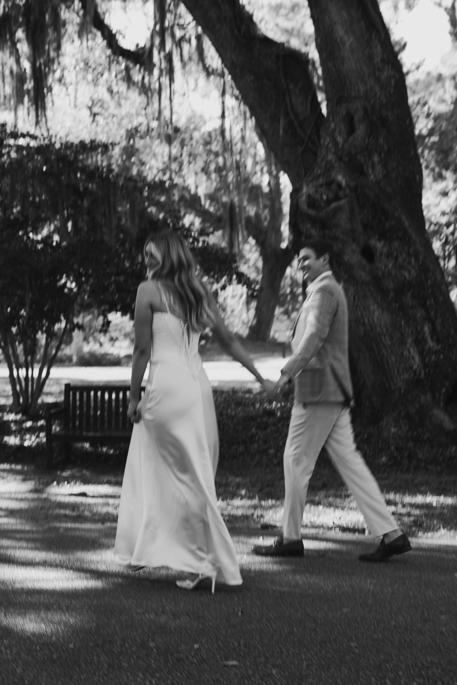 Black and white photo of a bride and groom walking hand in hand through a park with Spanish moss trees.