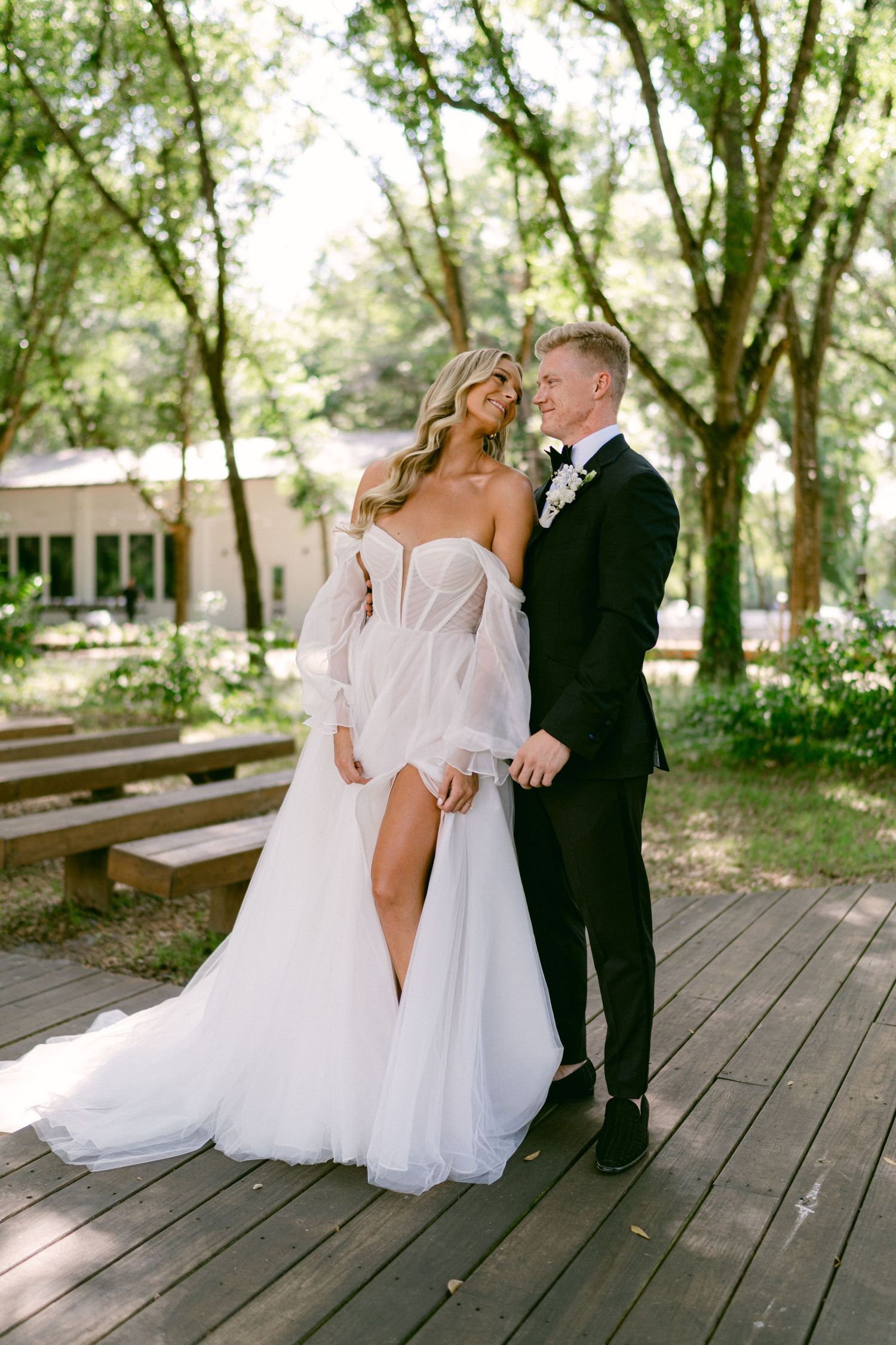 Bride in off-shoulder tulle gown with slit and groom in black tuxedo embracing on wooden deck surrounded by trees.