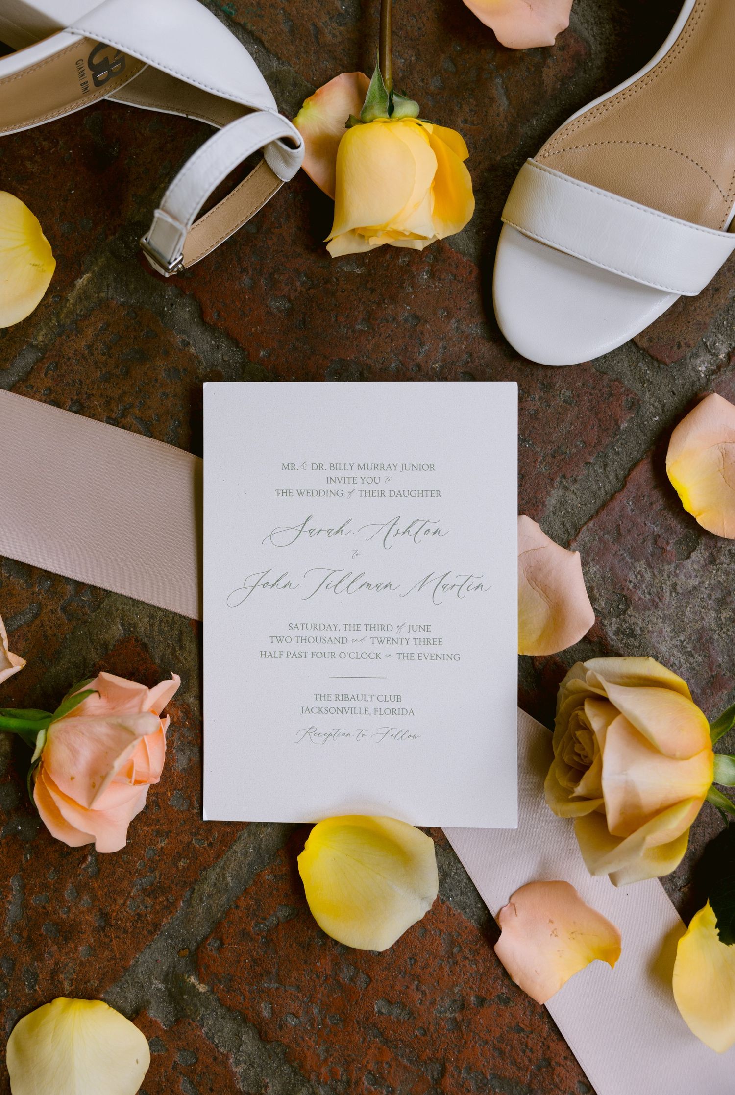 Elegant wedding invitation surrounded by yellow and pink roses and white heels on a marble surface.