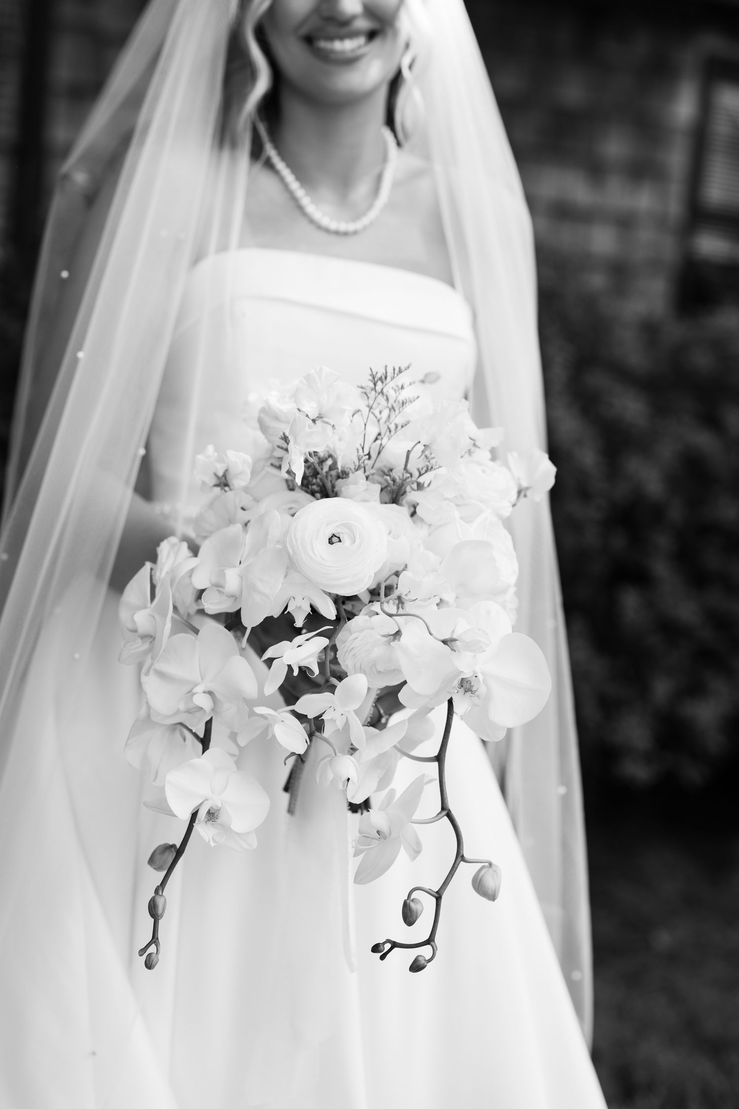 Bride in white strapless gown and veil holds cascading orchid and ranunculus bouquet in elegant black and white photo.