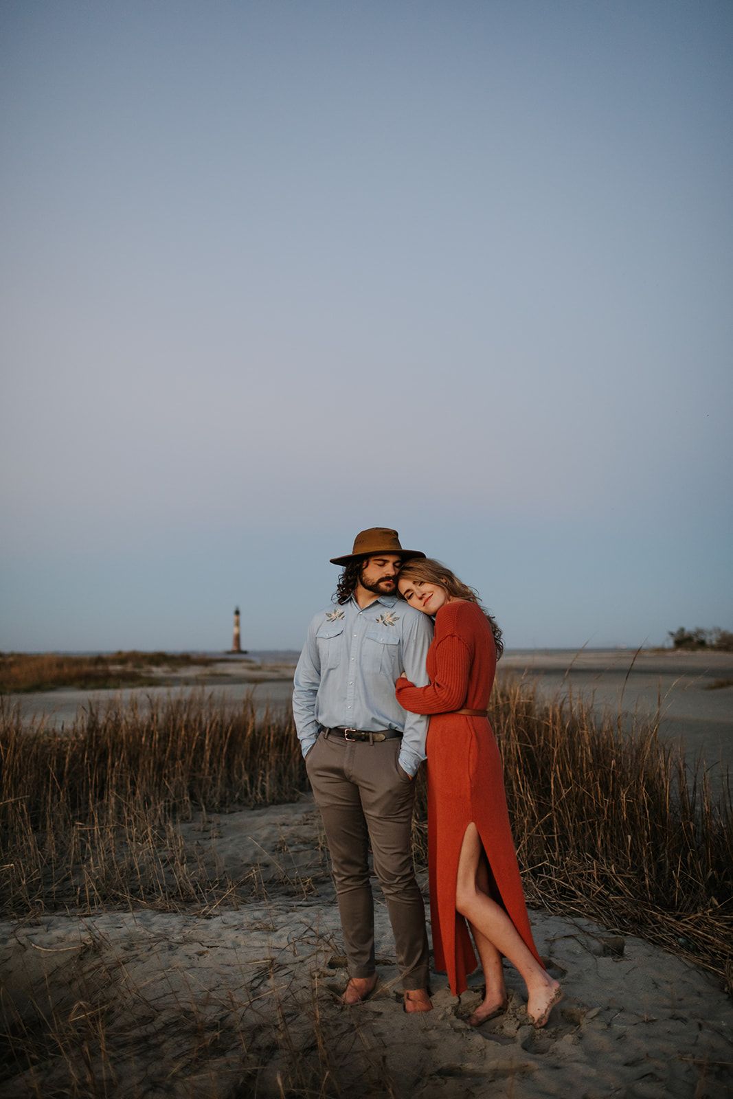 Couple poses at golden hour on sandy beach dunes, woman in red dress leaning on mans shoulder.