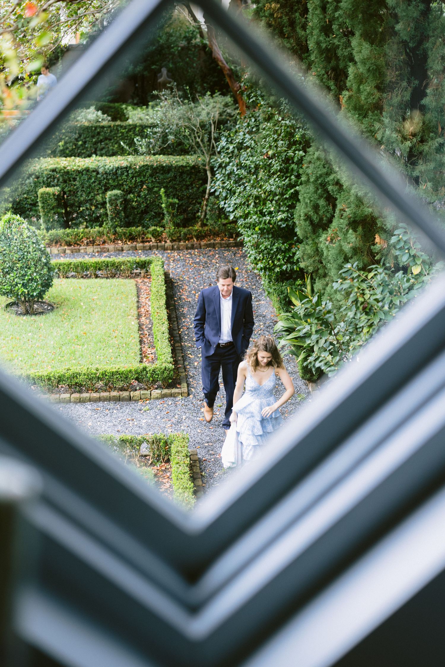 Couple walking through formal garden viewed through diamond-shaped window pane from above.