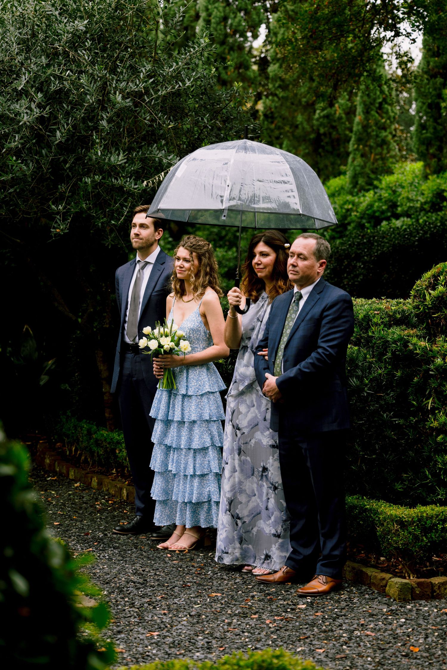 Wedding guests standing under clear umbrella in garden during rain, bridesmaid in blue ruffled dress.