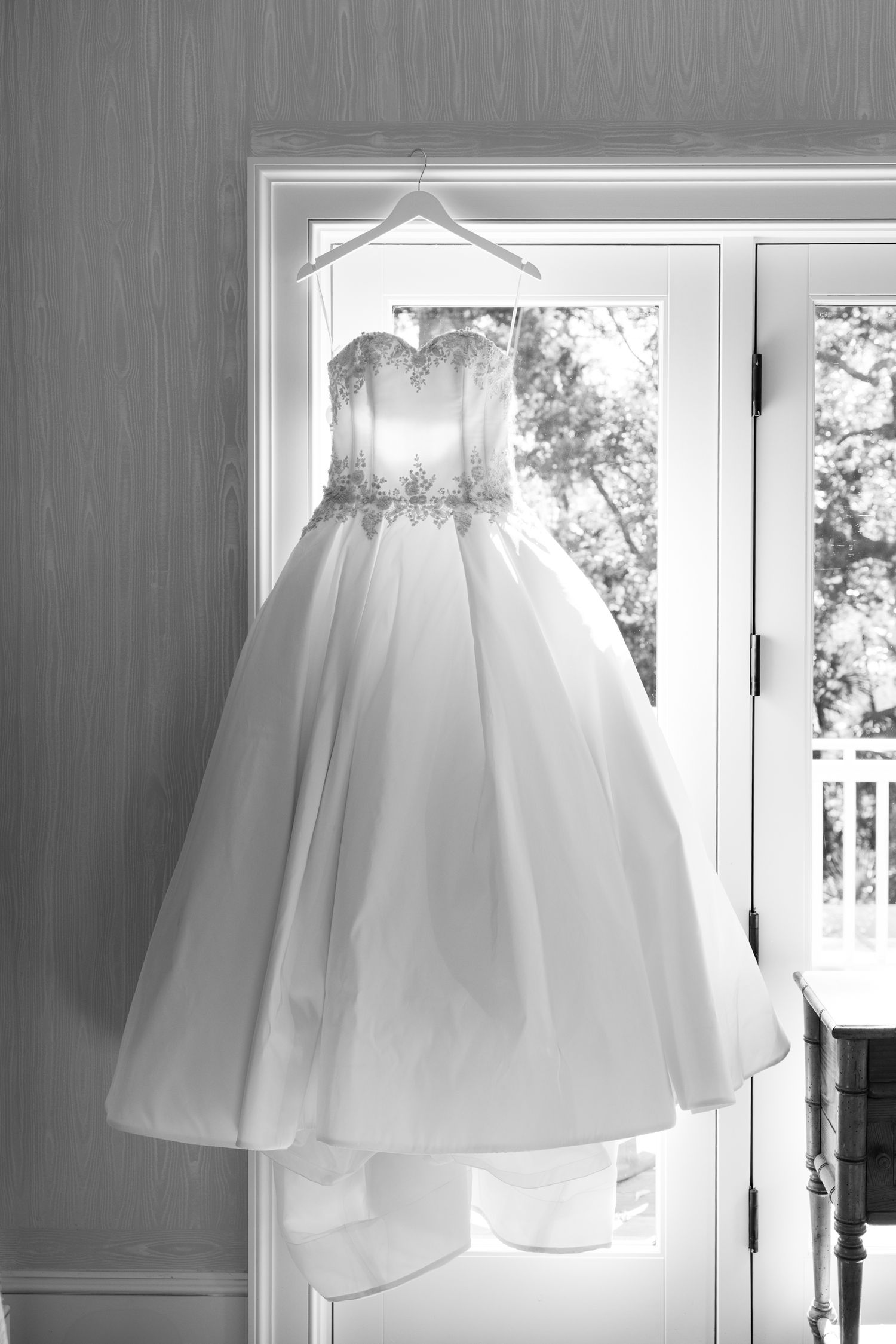 Black and white photo of an elegant strapless ball gown wedding dress with beaded bodice hanging by a sunlit window.