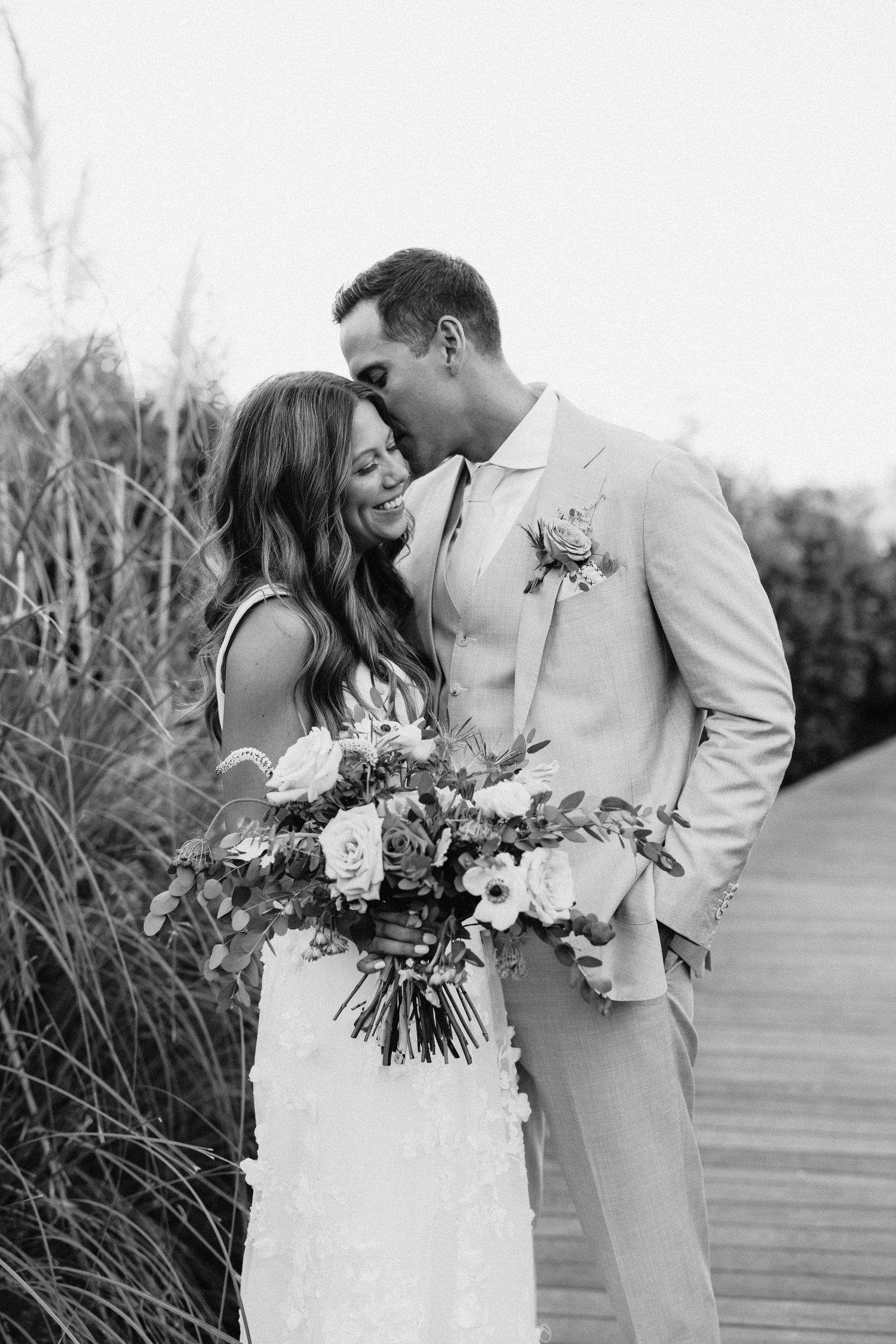 Black and white photo of a bride and groom sharing a tender moment on a wooden boardwalk, bride holding a lush floral bouquet.