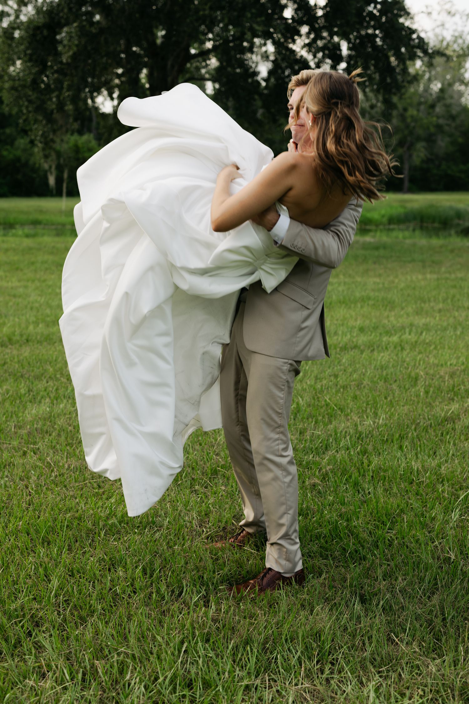 Groom in tan suit lifting bride in white wedding dress outdoors on green grass with trees in background.