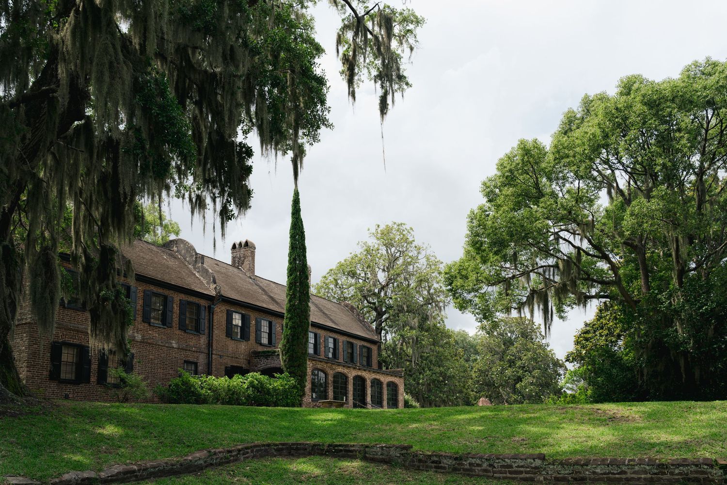 Elegant historic brick plantation house framed by towering moss-covered oak trees on a cloudy day.
