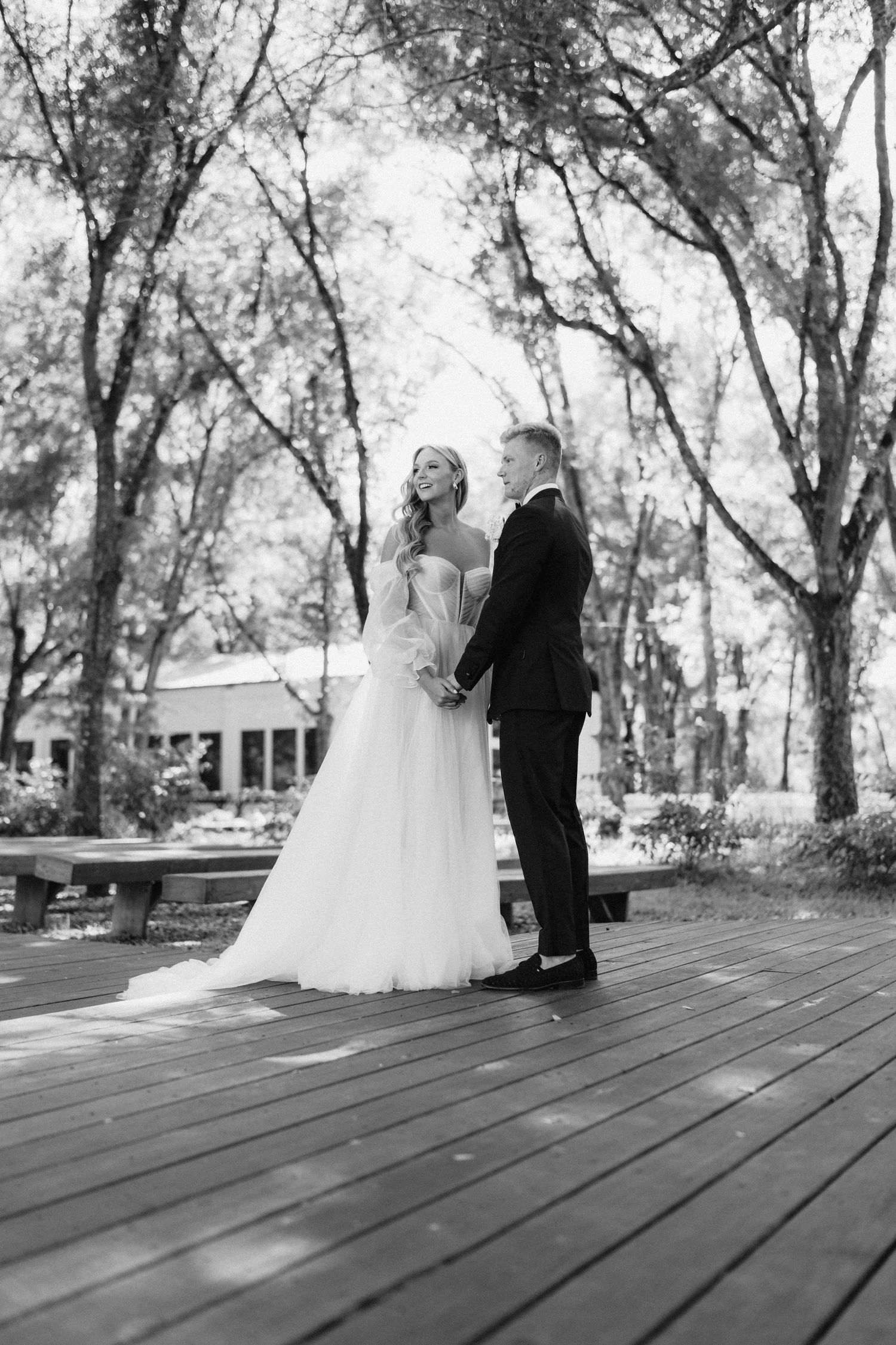 Black and white photo of bride and groom holding hands on wooden deck surrounded by tall trees.