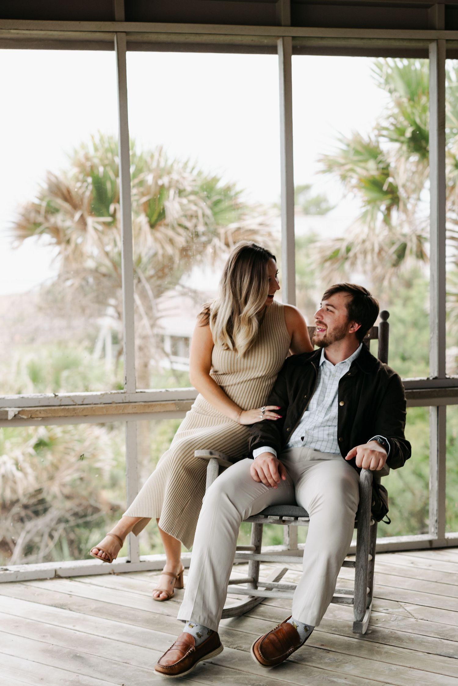 Couple sharing a romantic moment on a rustic porch with rocking chair, palm trees visible through large windows.