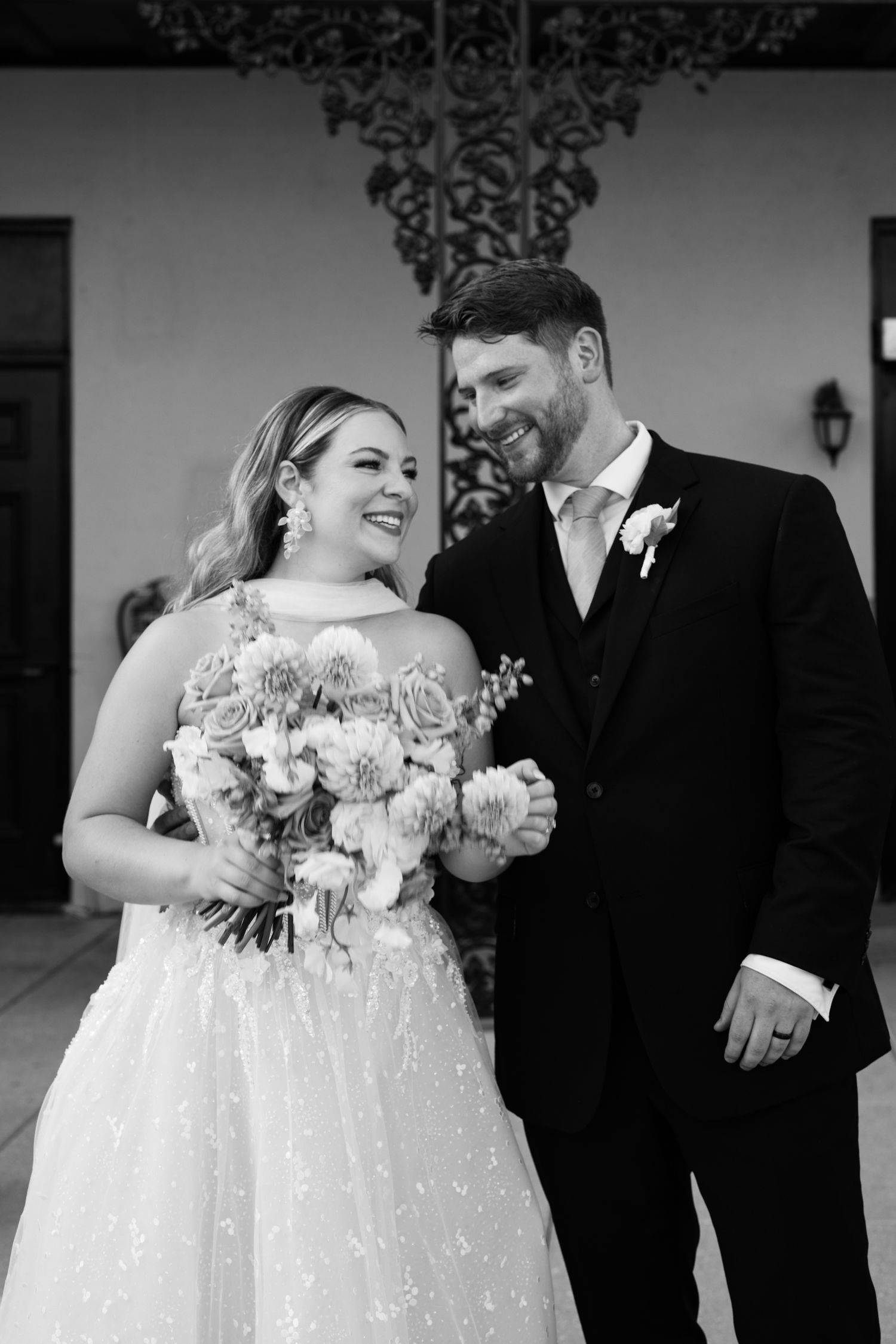 Black and white photo of a smiling bride holding a bouquet standing beside a groom in a suit at their wedding.