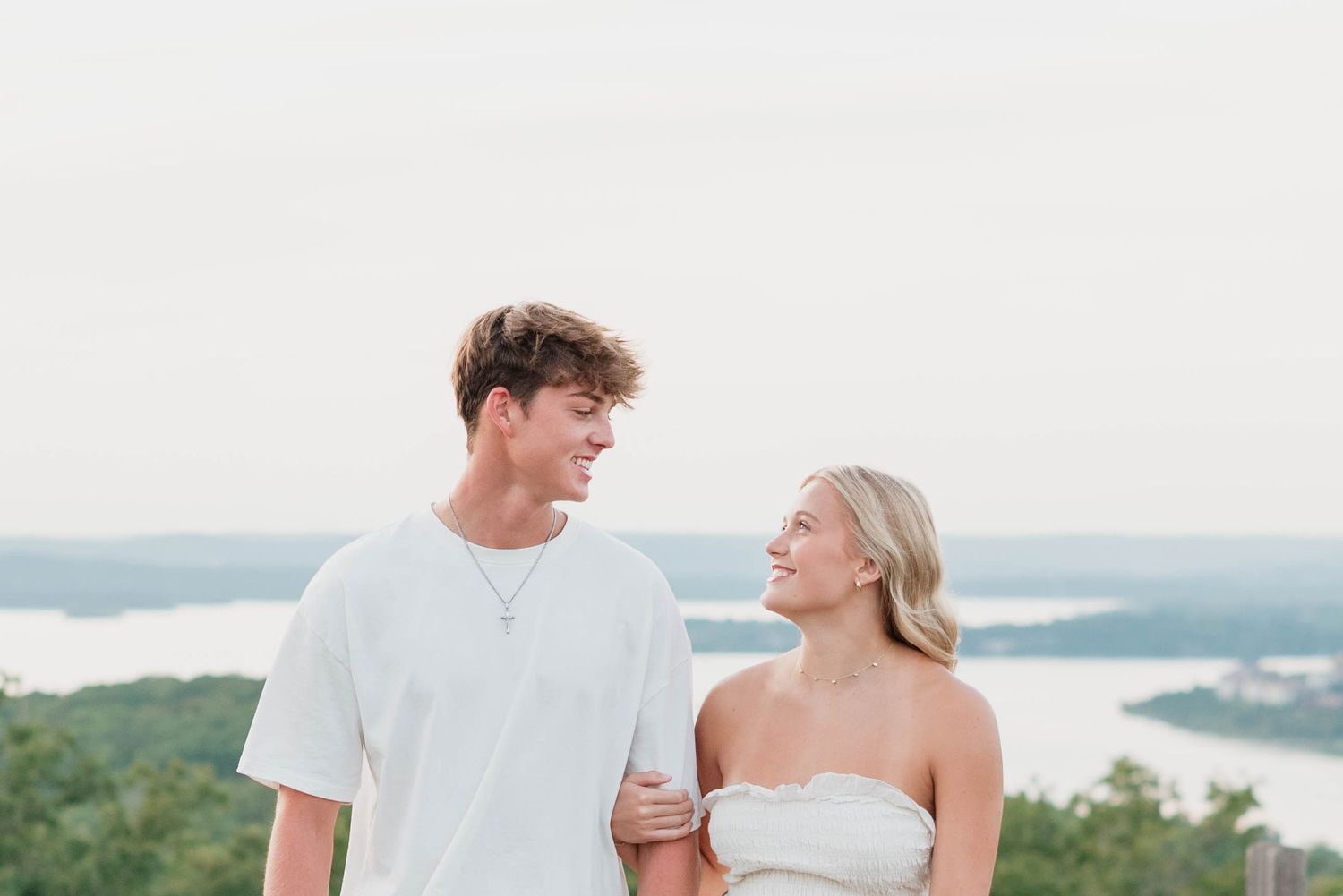 Young couple smiling at each other outdoors with scenic lake and green hills in the background.
