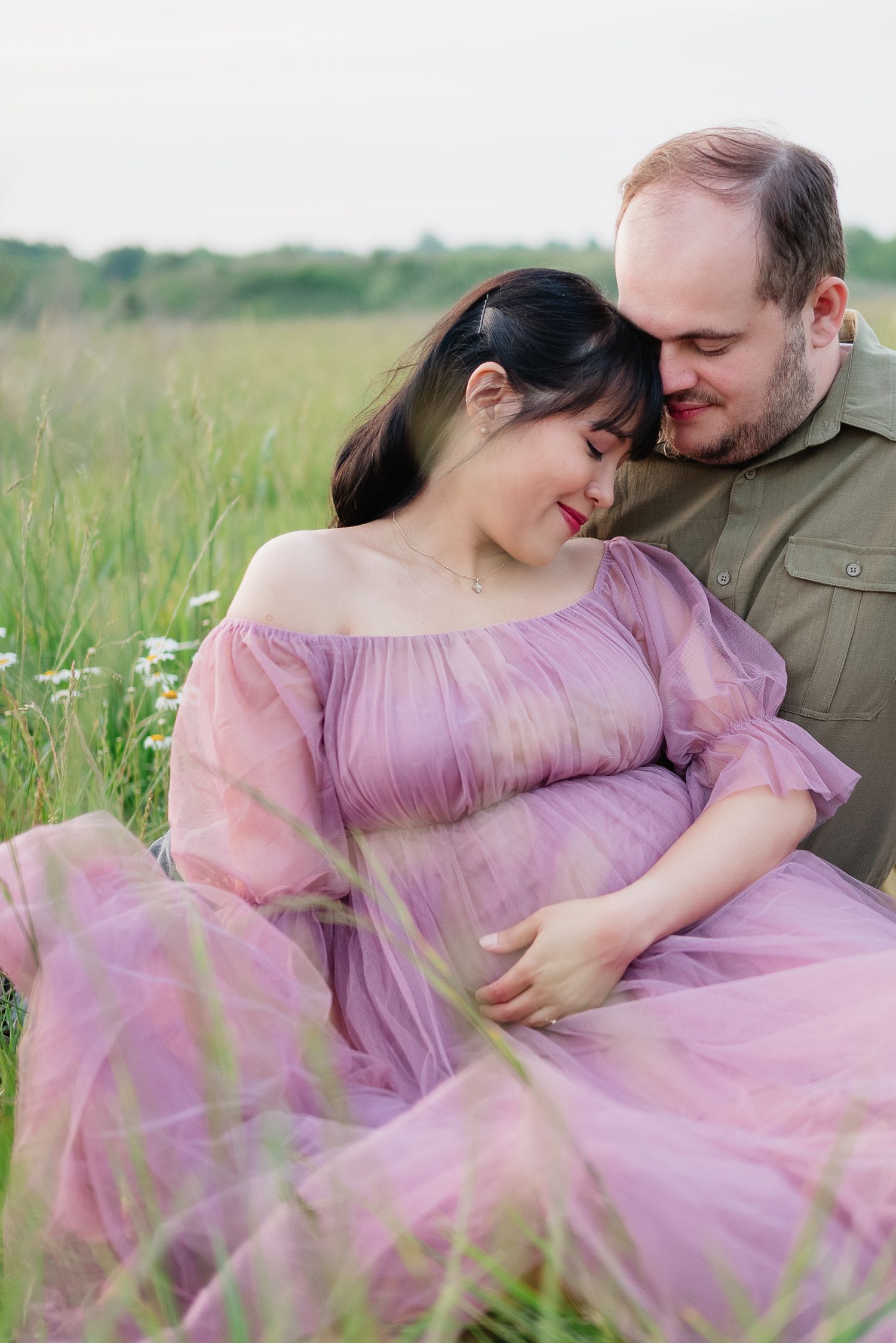 A couple sits in a green field, the pregnant woman in a pink dress leaning against her partner during a maternity photoshoot.