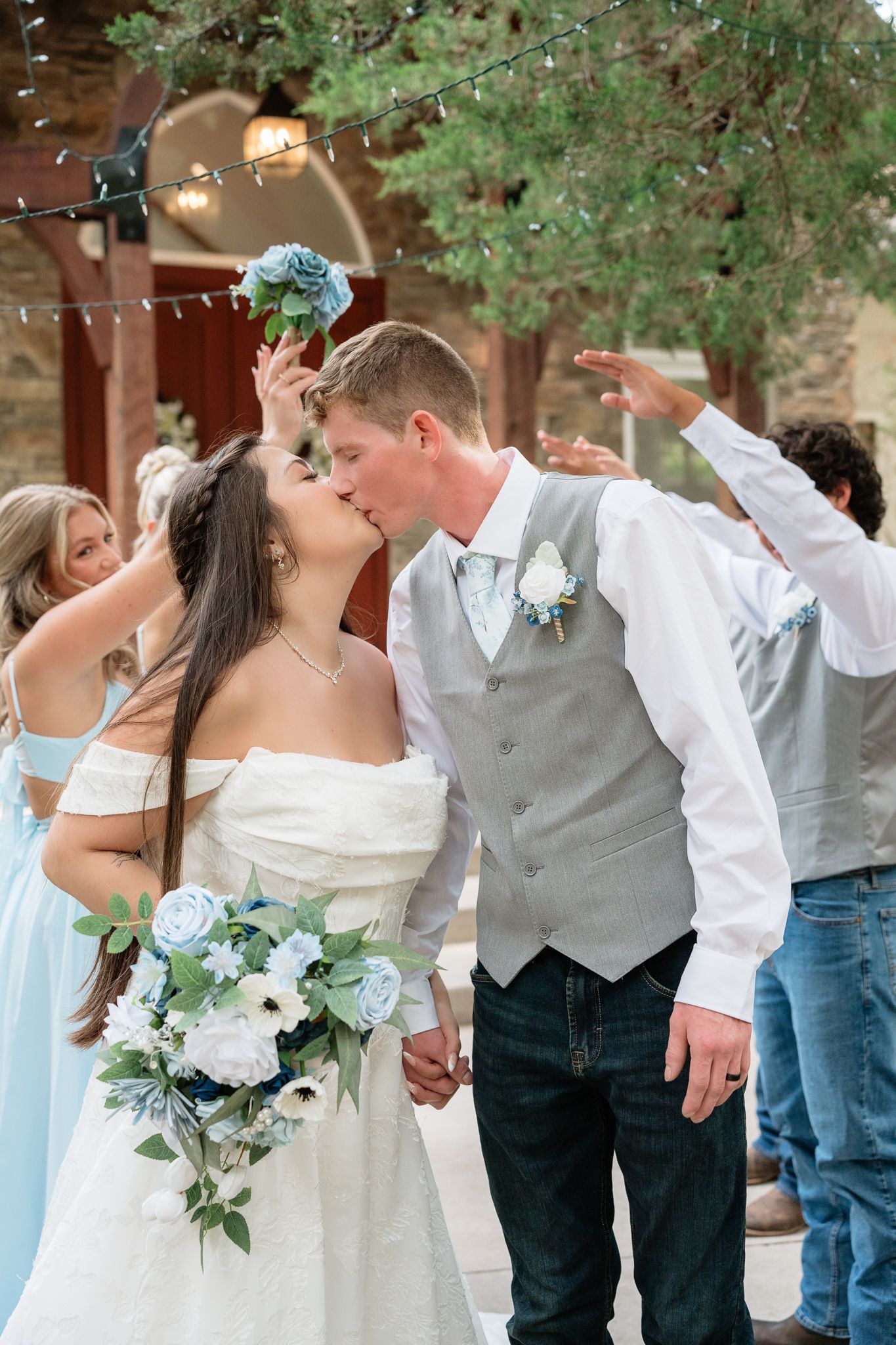 Bride and groom share a kiss holding blue and white bouquet outdoors at rustic wedding venue with string lights.
