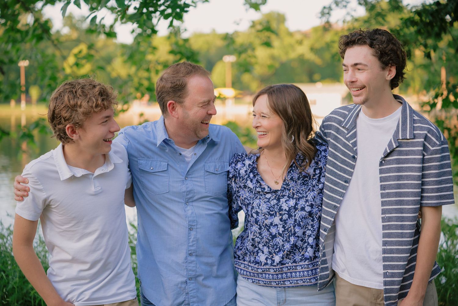 Family of four laughing together outdoors near a pond, enjoying a warm summer day.