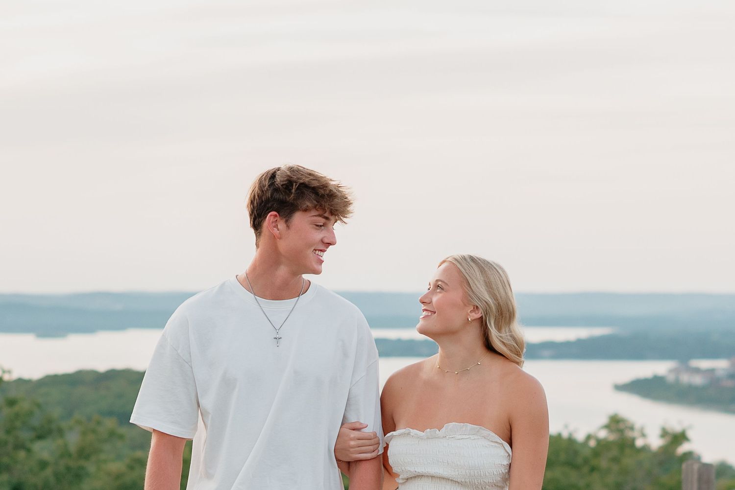 A young couple smiles at each other outdoors, with a scenic lake and lush green hills in the background.