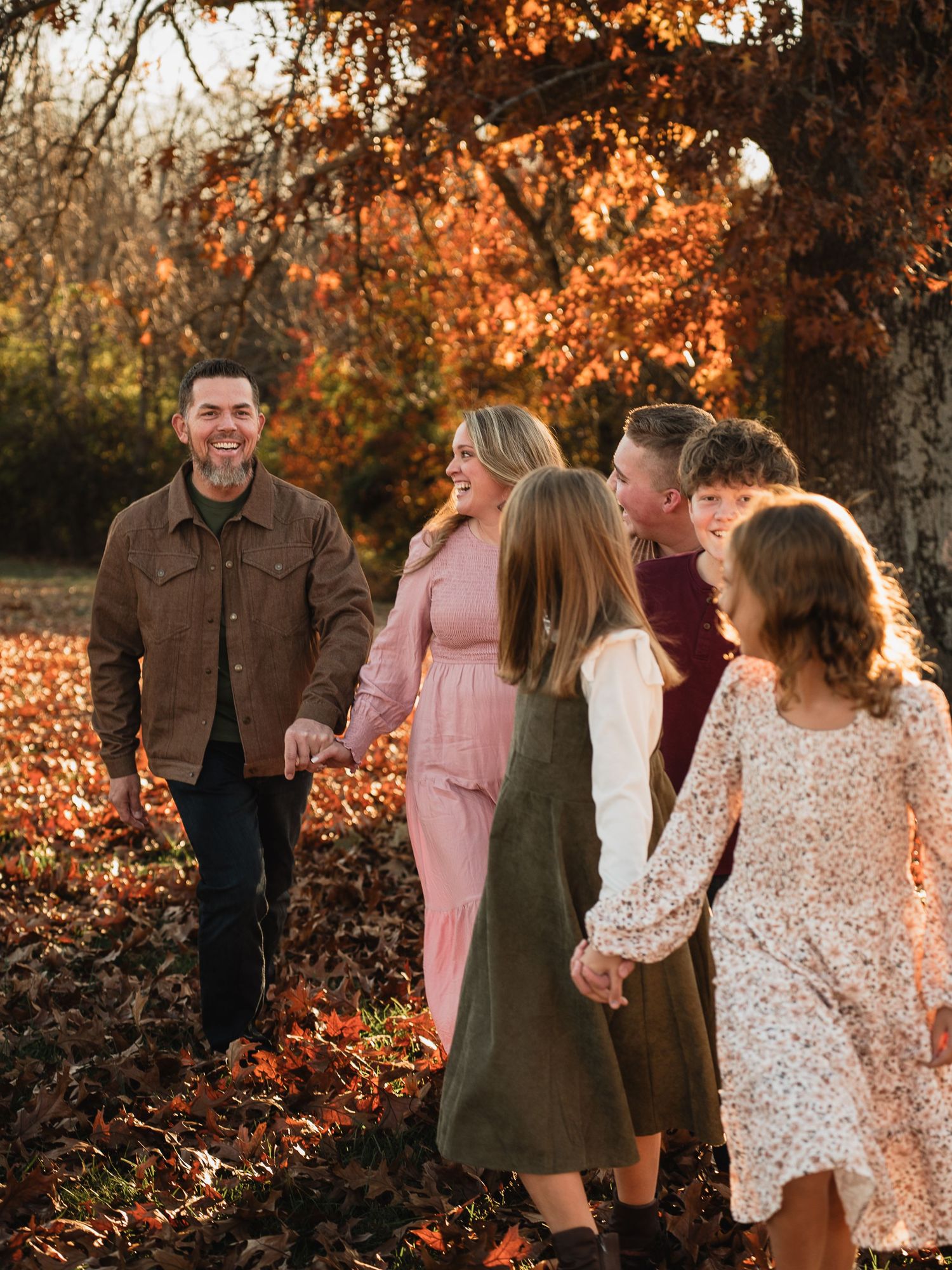 Family walking through autumn leaves, laughing together in golden fall light among colorful orange and red trees.