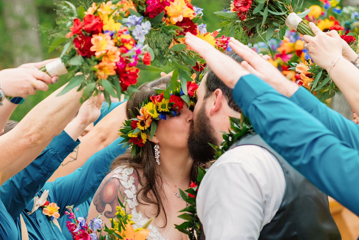 Bride and groom kiss under an arch of colorful bouquets held by bridesmaids in teal dresses at outdoor wedding.
