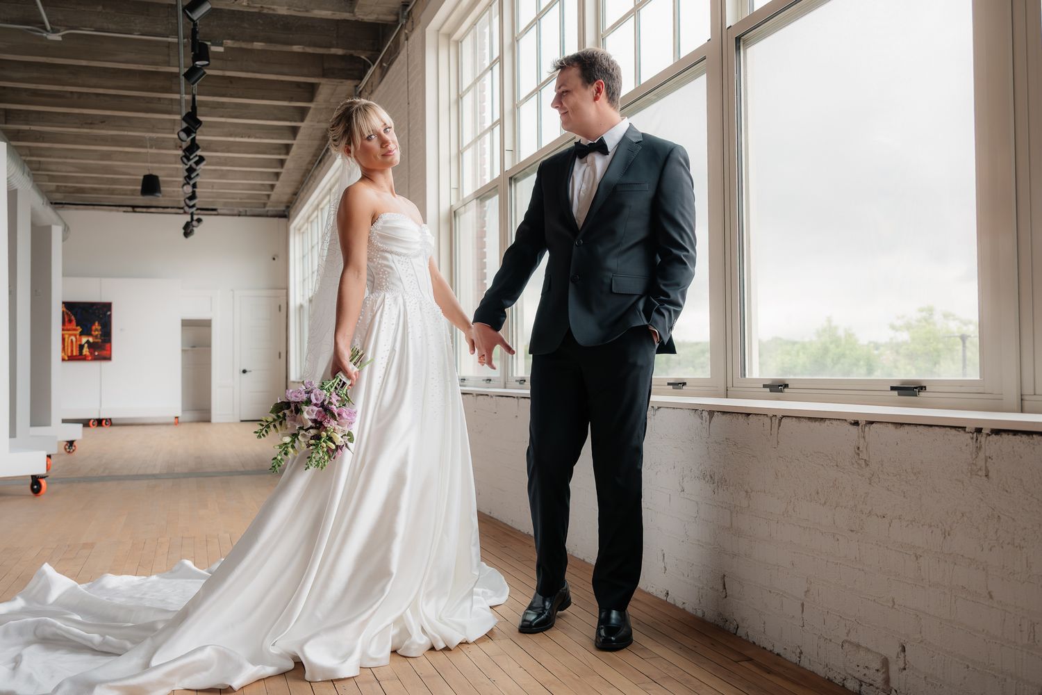 Bride in white gown and groom in black tuxedo hold hands by large windows in elegant industrial venue.