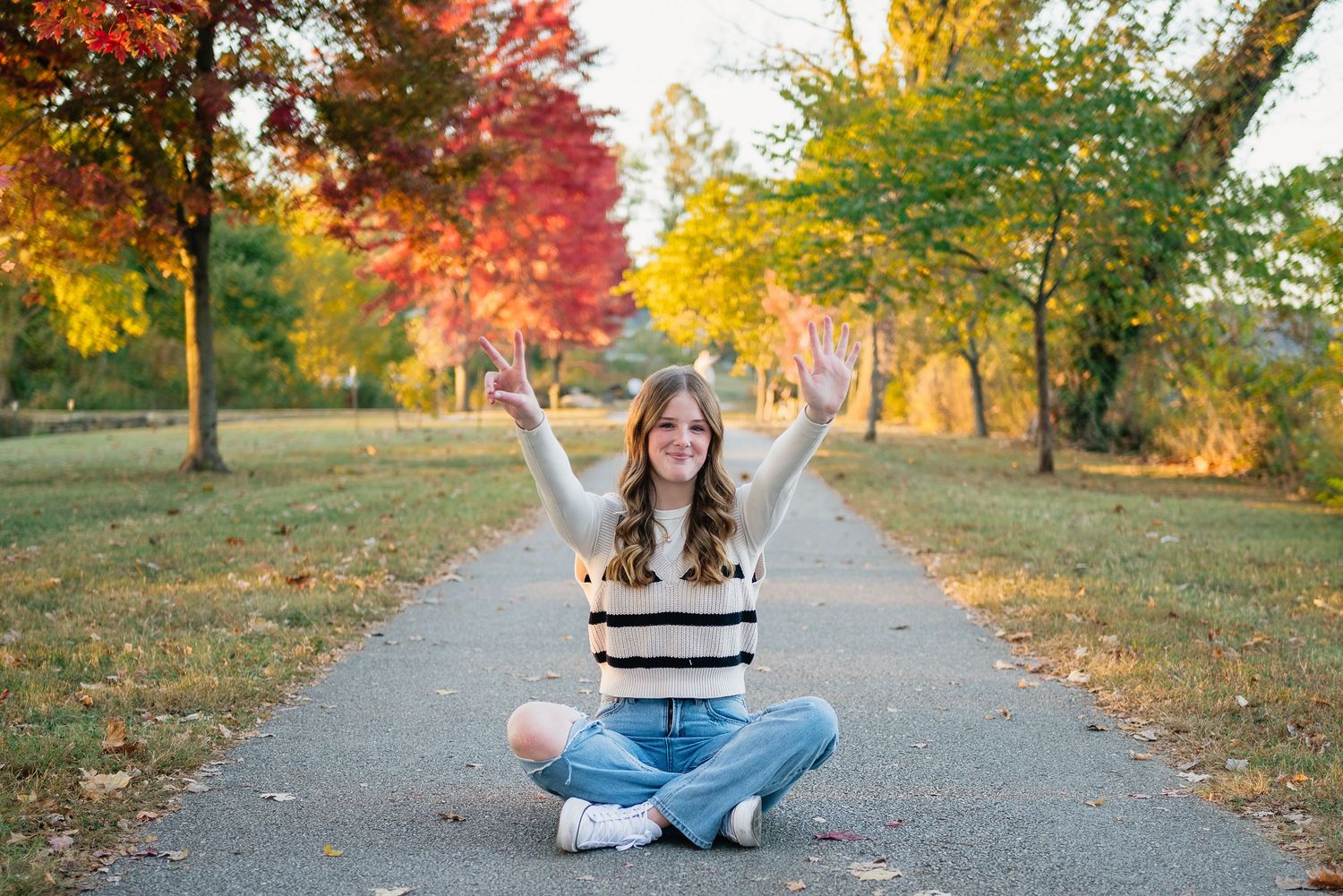 Young woman sitting cross-legged on a park path, arms raised joyfully, surrounded by colorful autumn trees.