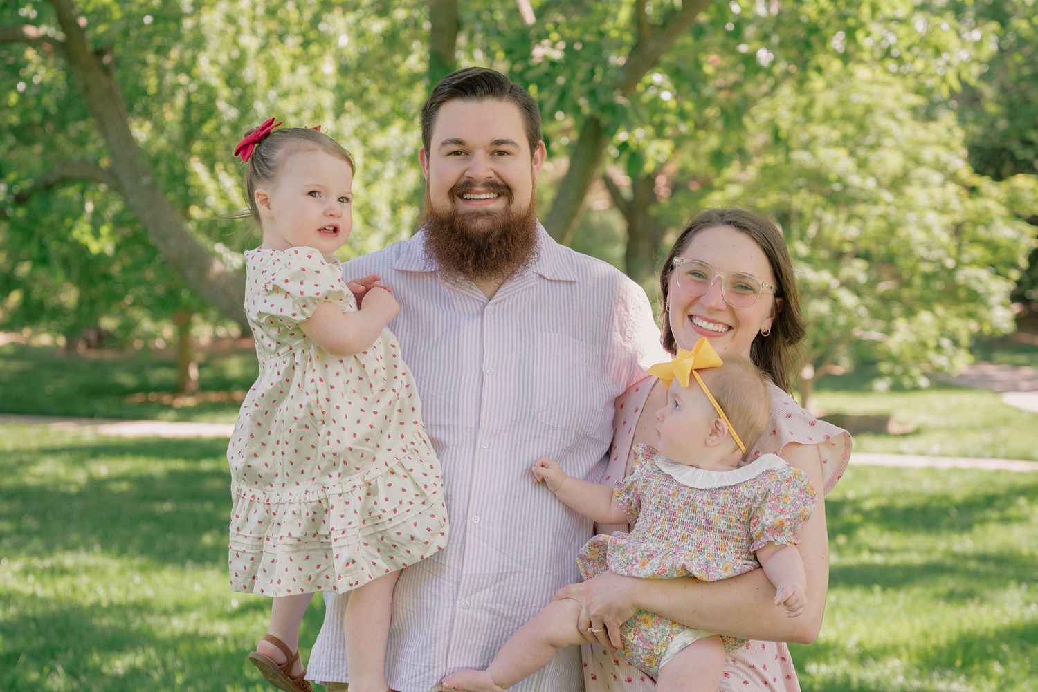Happy family with bearded dad, smiling mom, and baby girl in floral dress posing outdoors in a lush green park.